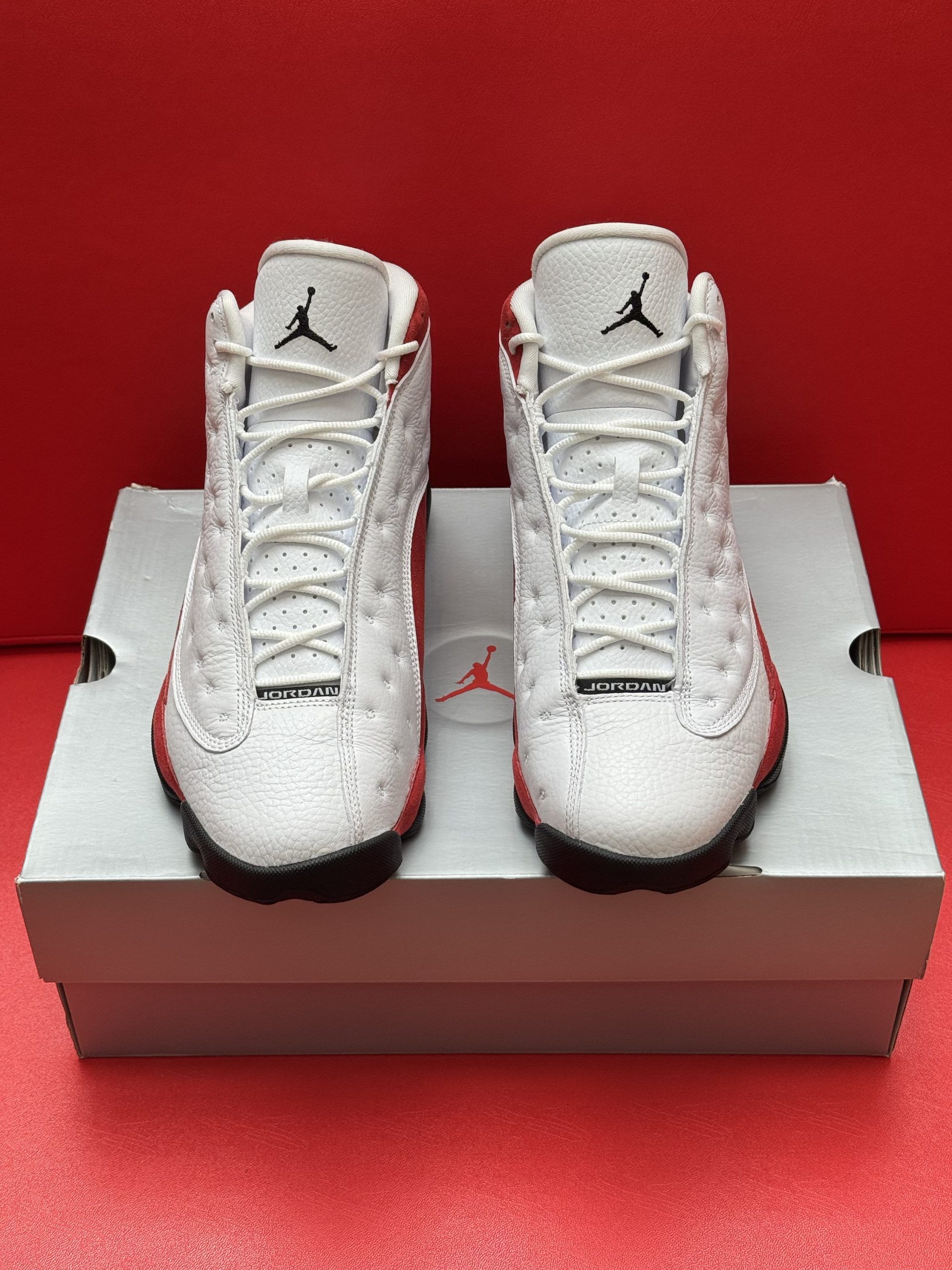 White and red Air Jordan 13 toddler sneakers on a box, red background.