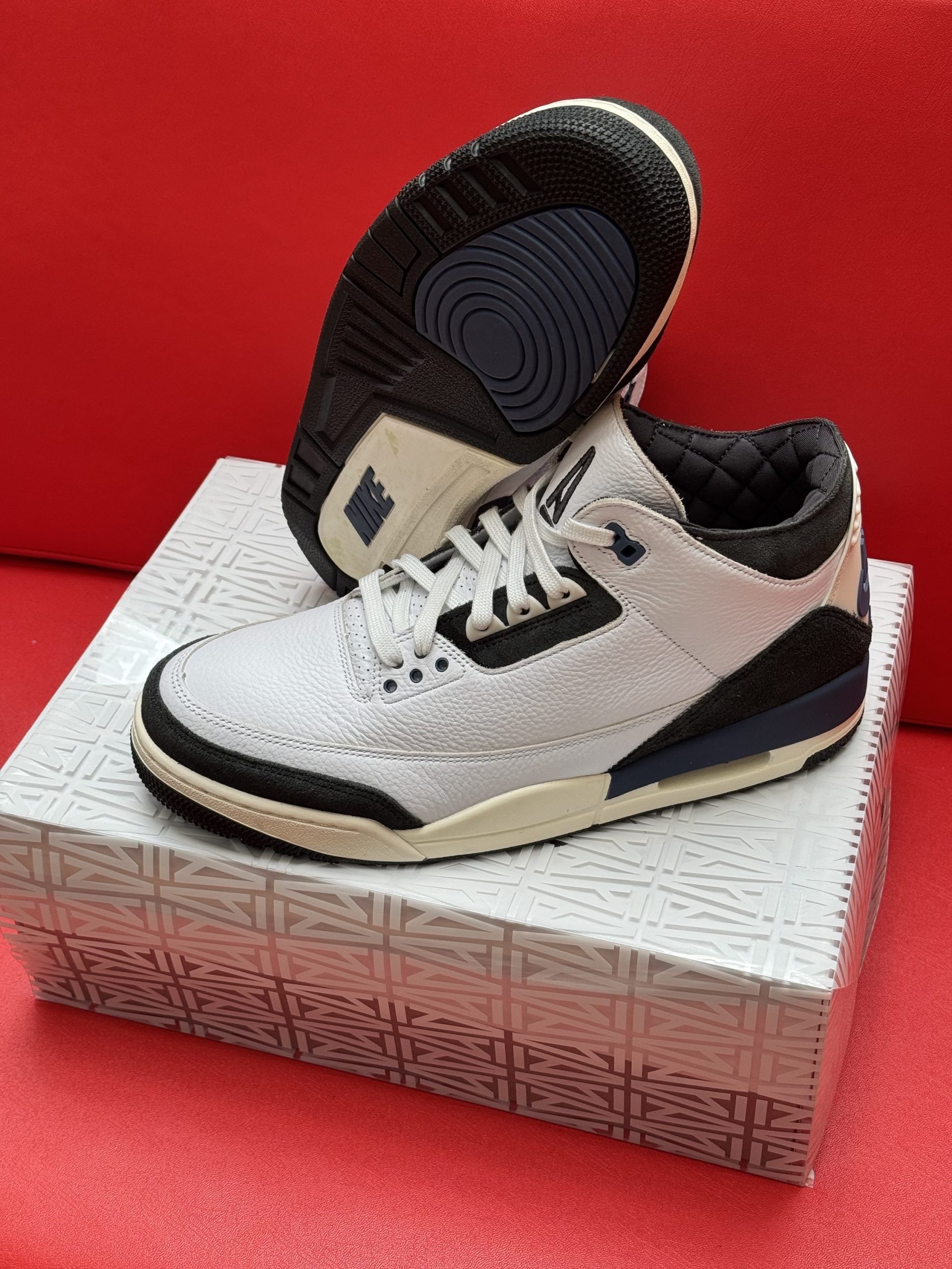 White and black Air Jordan 3 sneakers on a patterned box, against a red background.