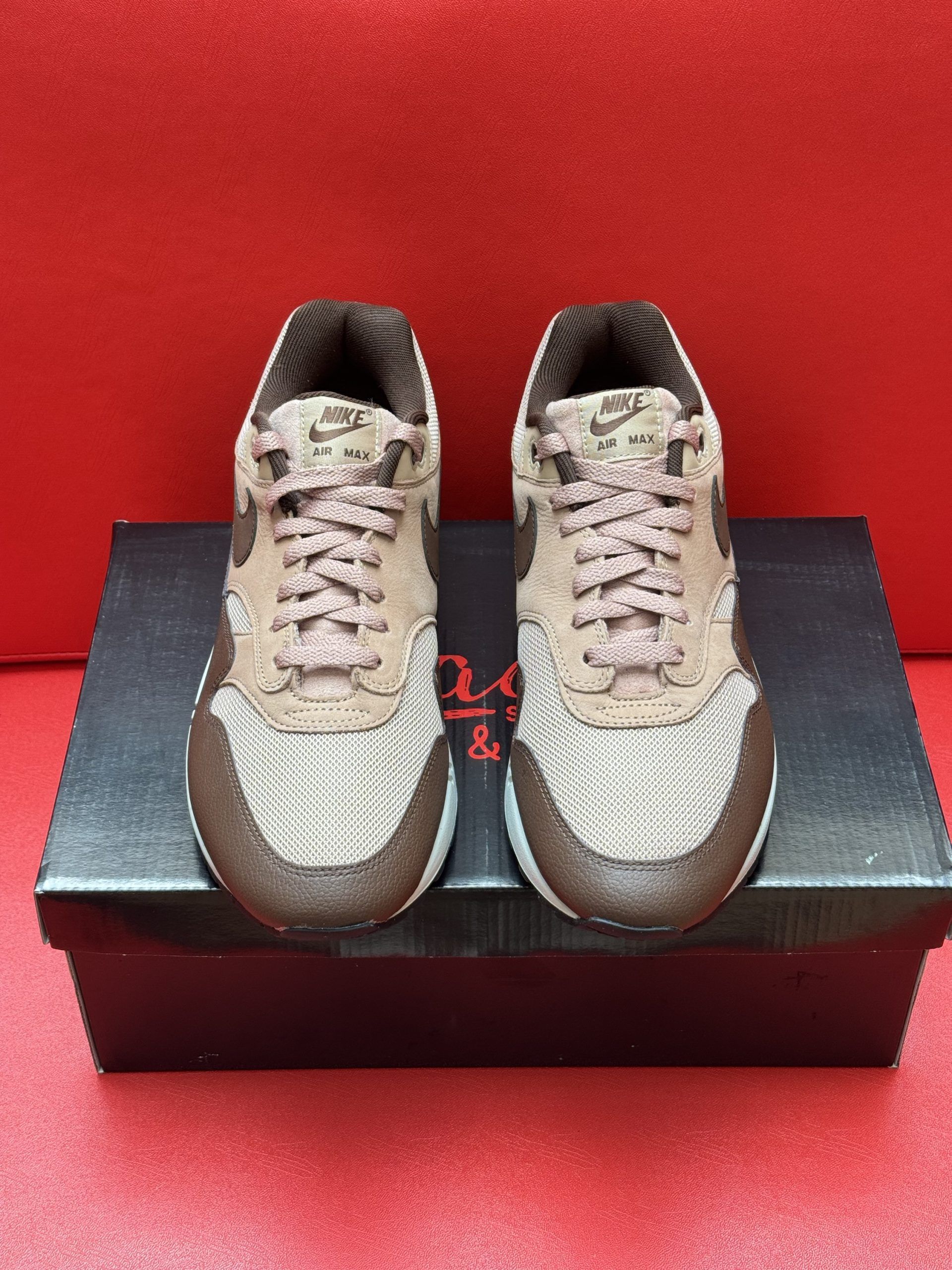 Brown and beige Nike Air Max 1 sneakers on a black shoebox, with the sole visible.