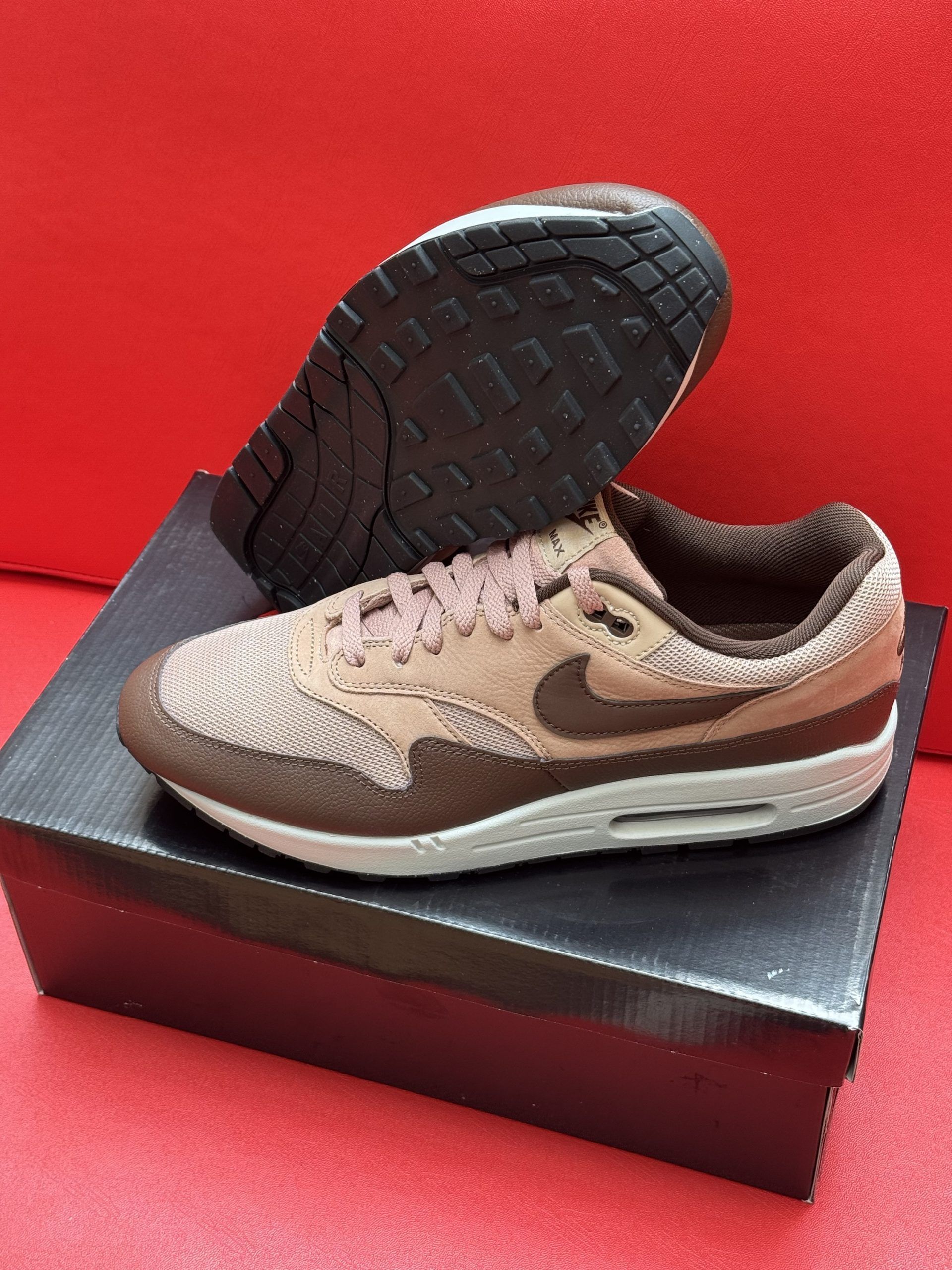 Brown and beige Nike Air Max 1 sneakers on a black shoebox, with the sole visible.