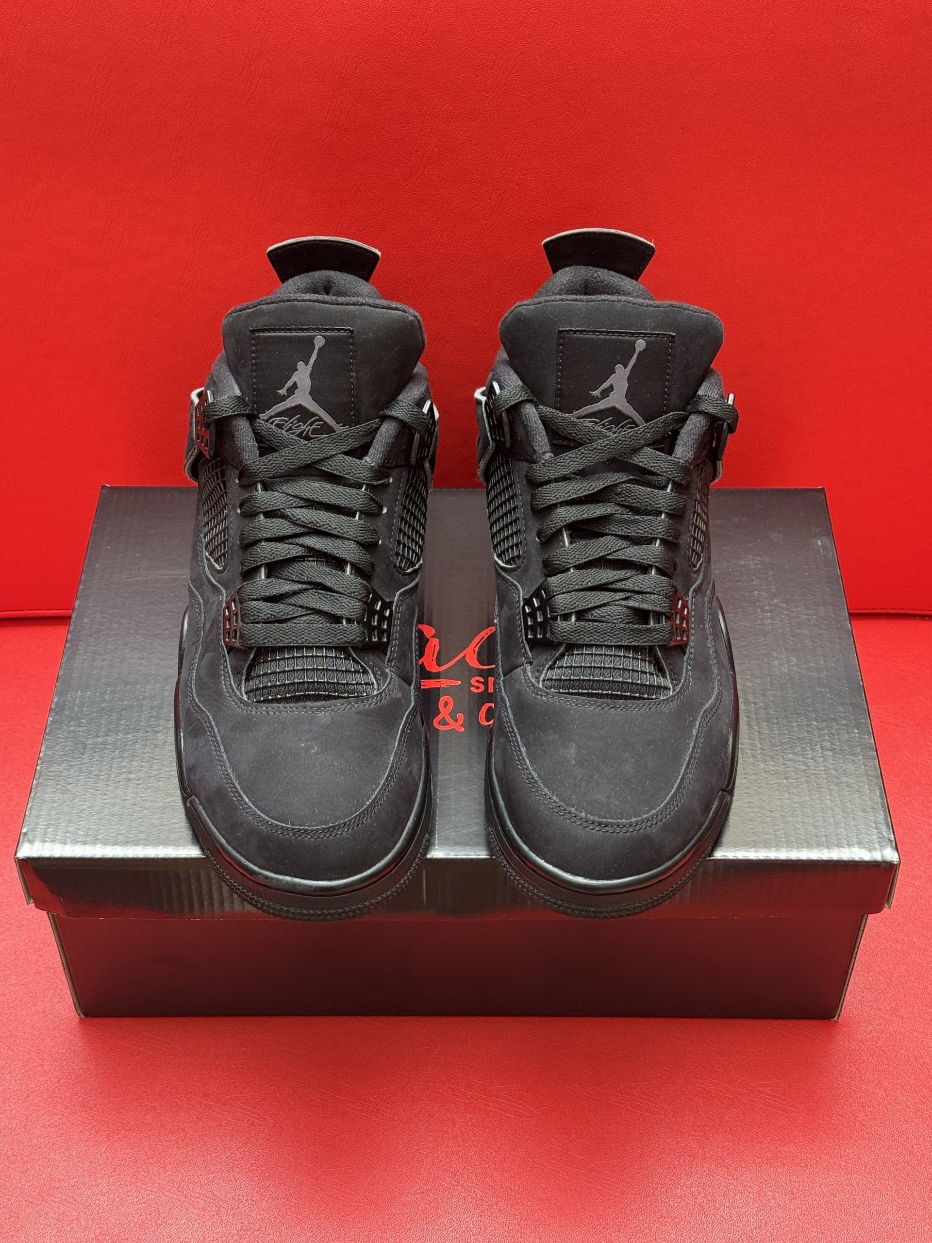 Black Jordan 4 baby shoes on a black box, set against a red background.
