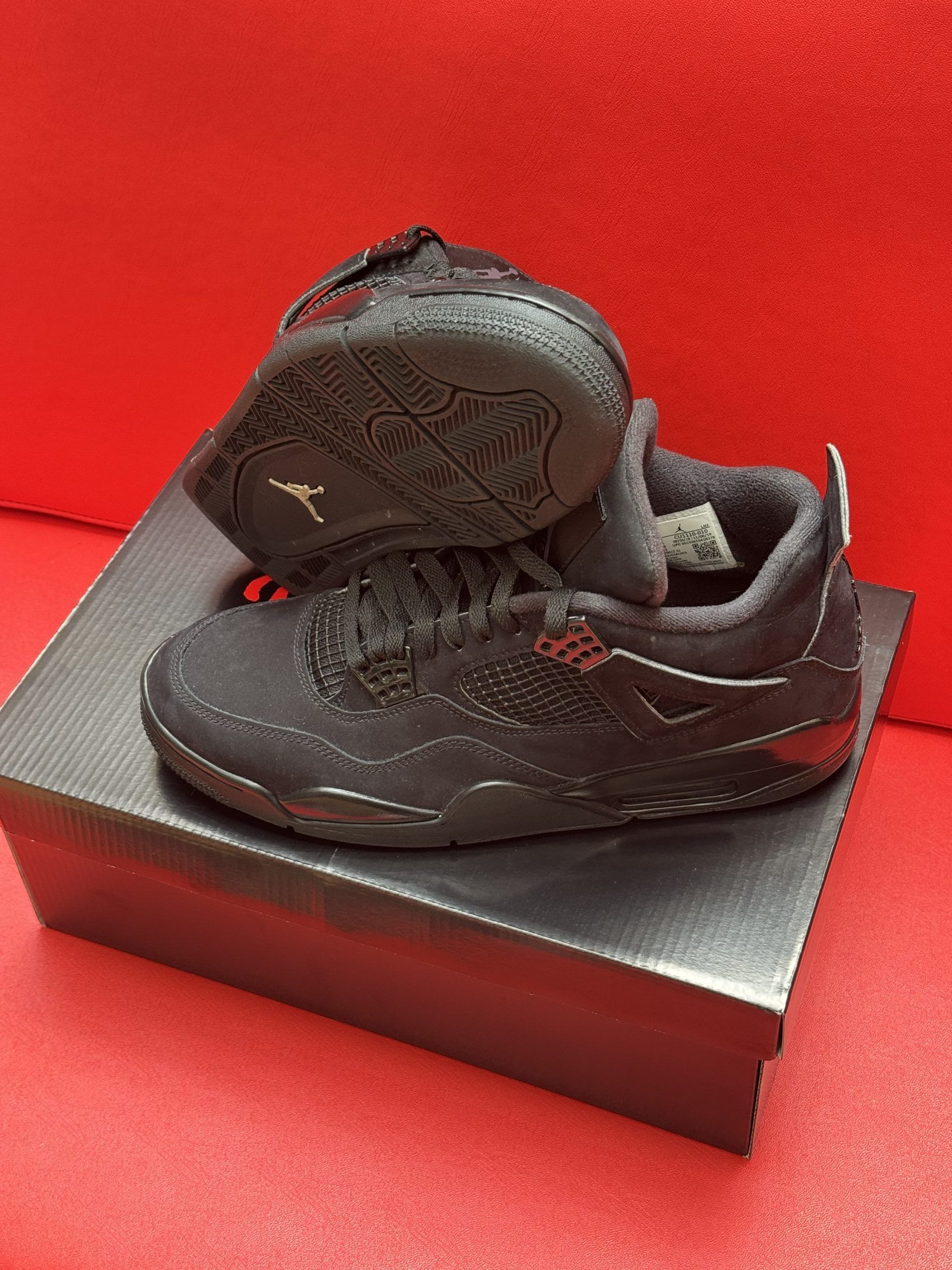 Black Jordan 4 baby shoes on a black box, set against a red background.