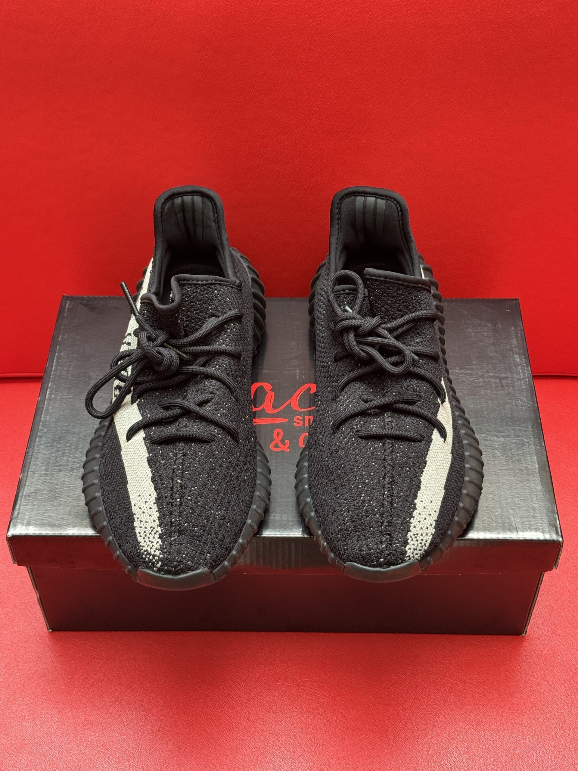 Black and beige Yeezy Boost 350 V2 sneakers on a black box, one shoe upside down showing the sole, against a red background.