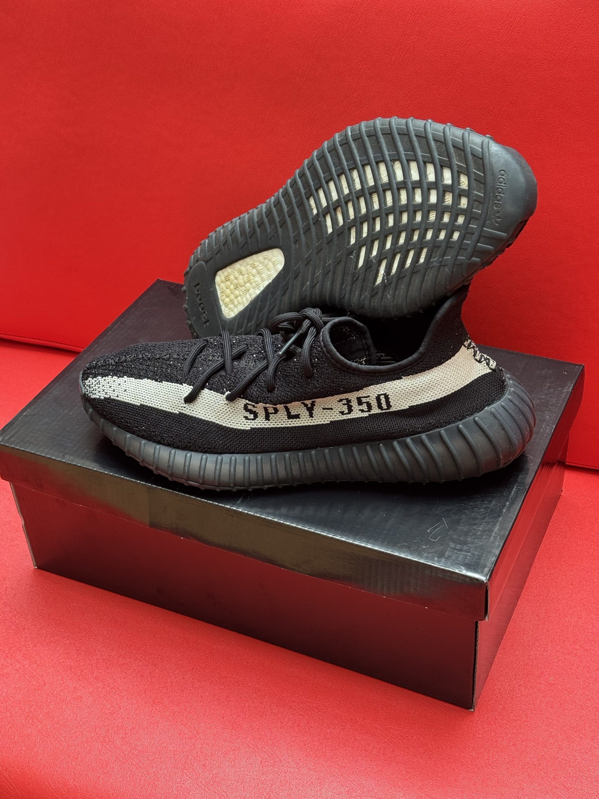 Black and beige Yeezy Boost 350 V2 sneakers on a black box, one shoe upside down showing the sole, against a red background.