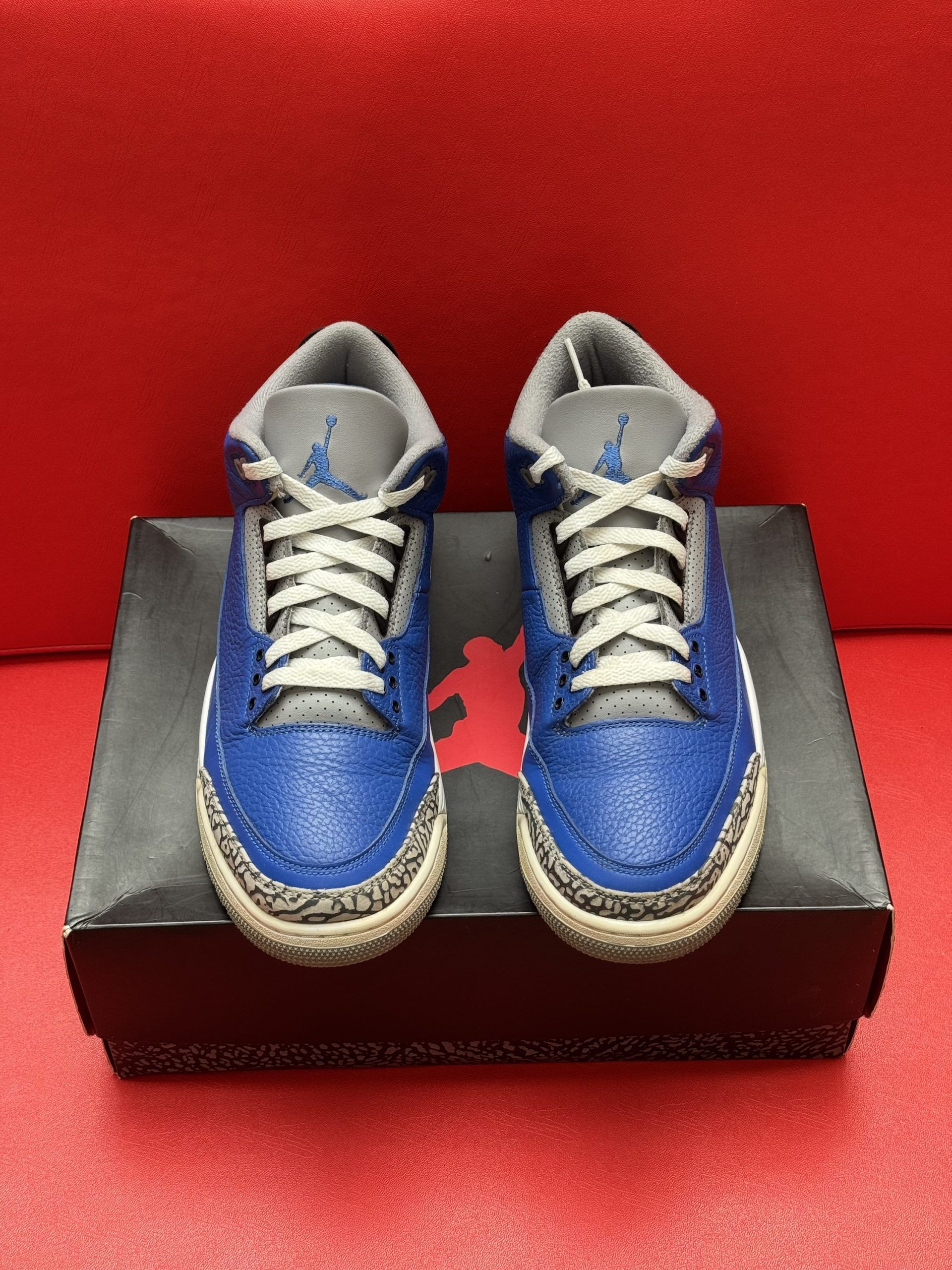 Blue and gray Air Jordan 3 sneakers on a black box, sitting on a red surface.