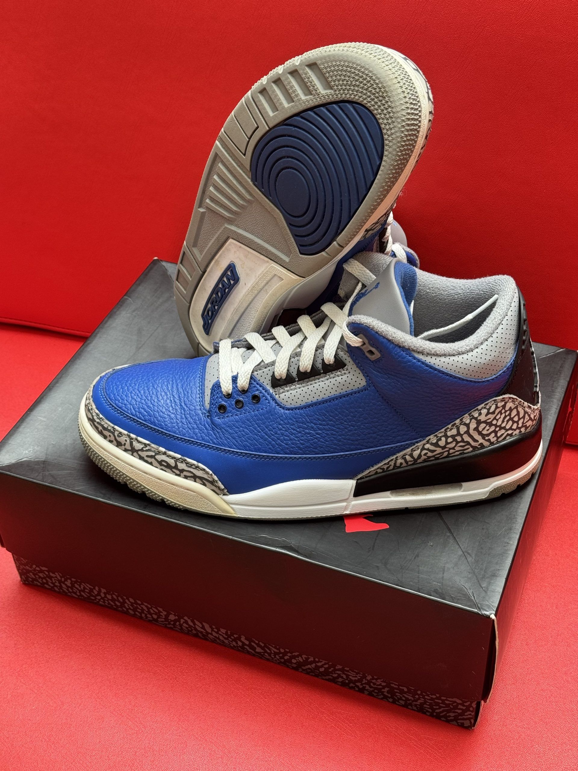 Blue and gray Air Jordan 3 sneakers on a black box, sitting on a red surface.