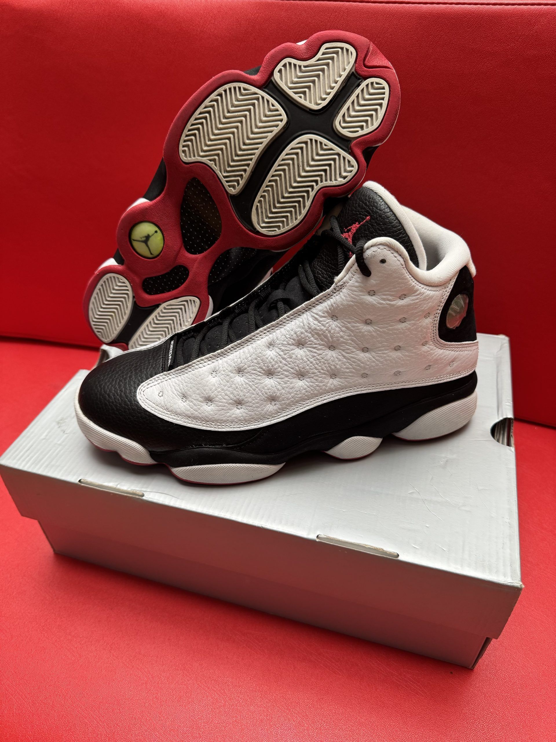 Air Jordan 13 sneakers, white and black with a red sole, resting on a shoebox against a red backdrop.