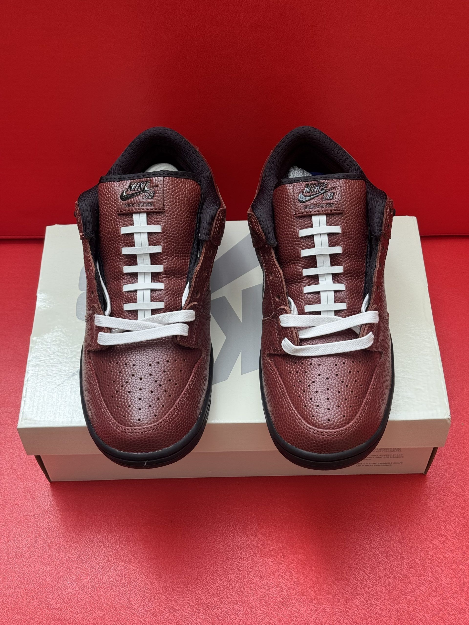 Nike Dunk Low sneakers, metallic brown with black sole and swoosh, white laces, sitting on a white box against a red backdrop.
