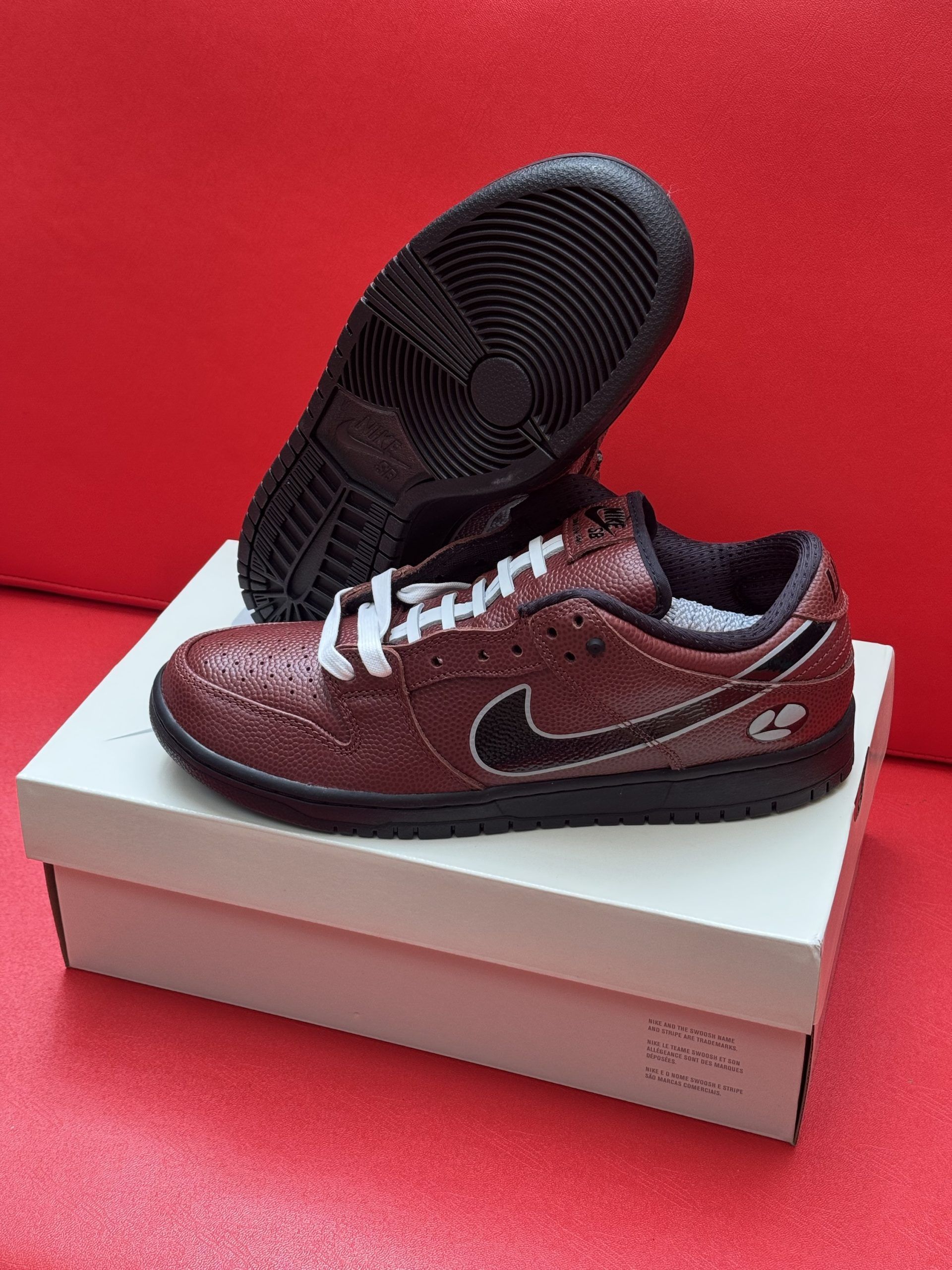 Nike Dunk Low sneakers, metallic brown with black sole and swoosh, white laces, sitting on a white box against a red backdrop.