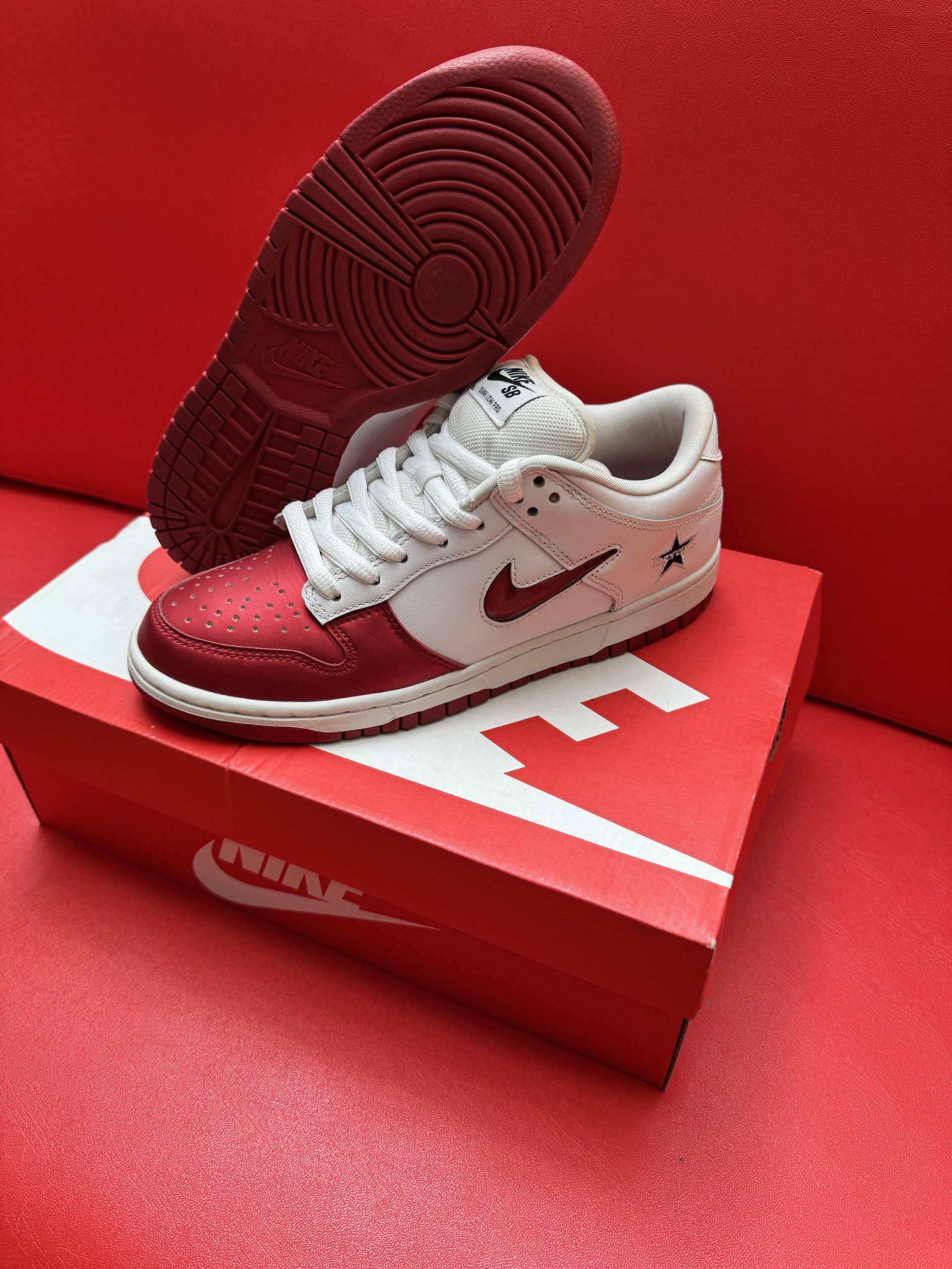 Red and white Nike SB Dunk Low sneakers with a star detail, on a red Nike box, against a red backdrop.