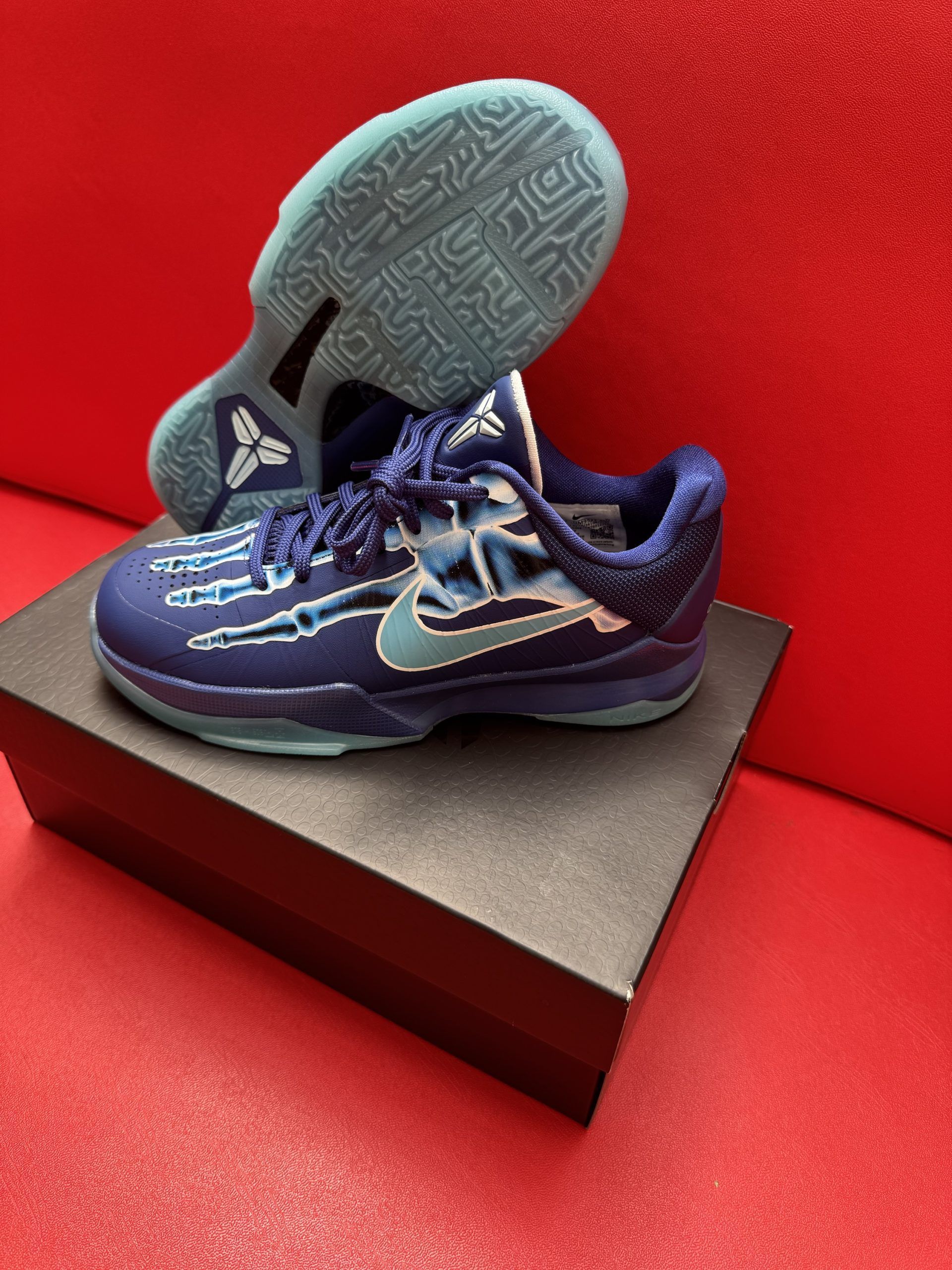 Blue and teal Nike Kobe basketball sneakers on a black box, sole visible. Red backdrop.