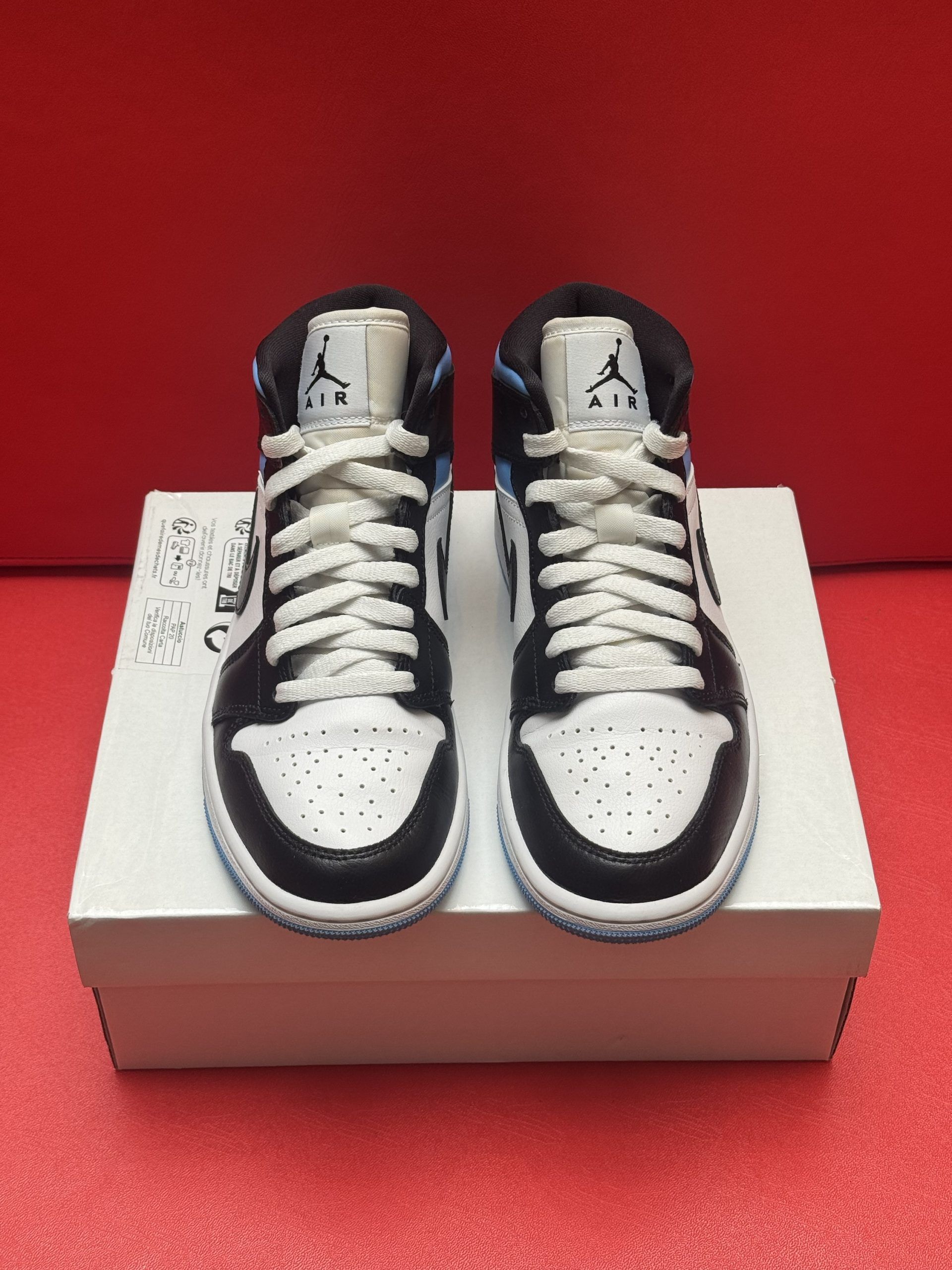 A pair of baby Air Jordan 1 sneakers, black, white, and light blue, on a white box against a red backdrop.