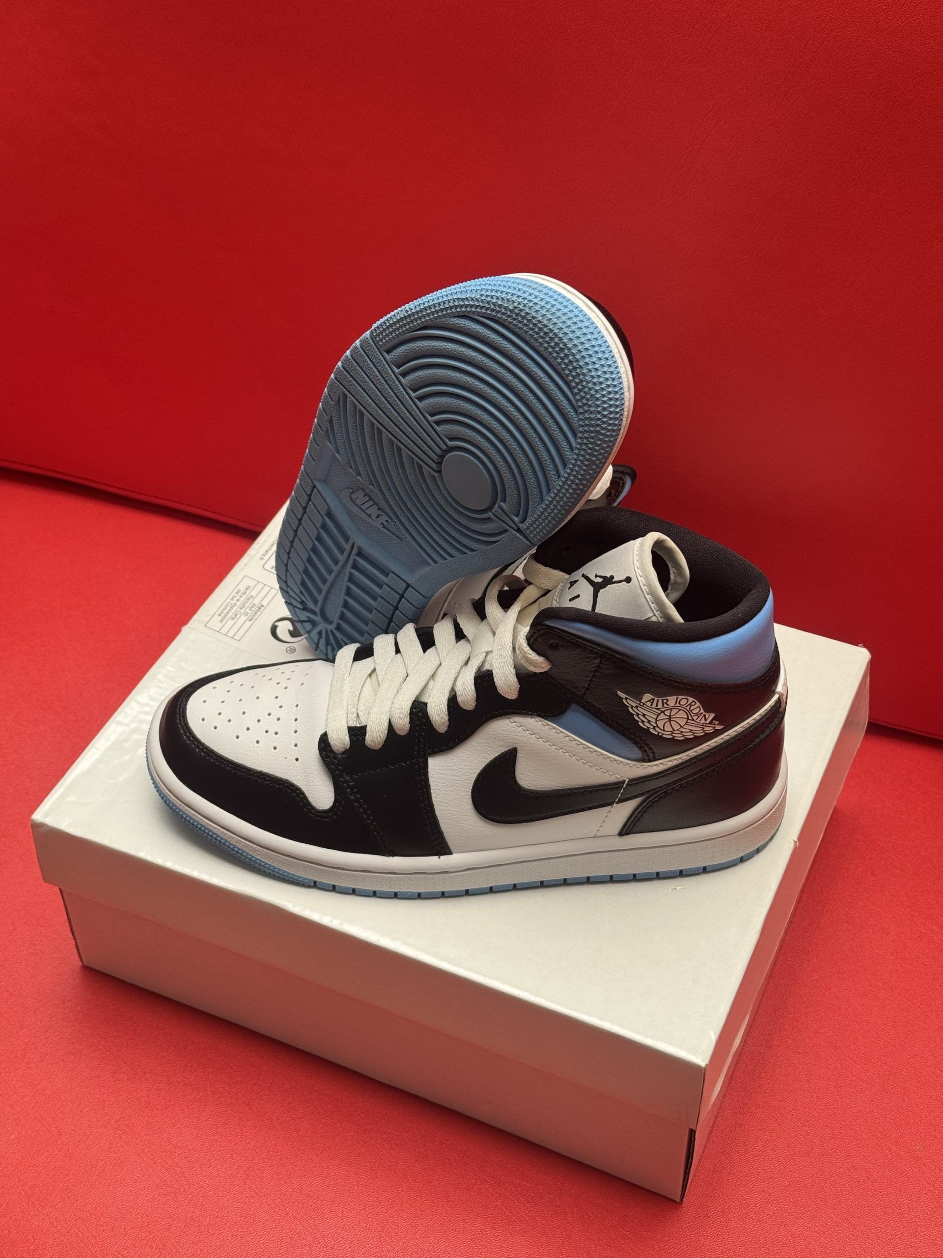 A pair of baby Air Jordan 1 sneakers, black, white, and light blue, on a white box against a red backdrop.