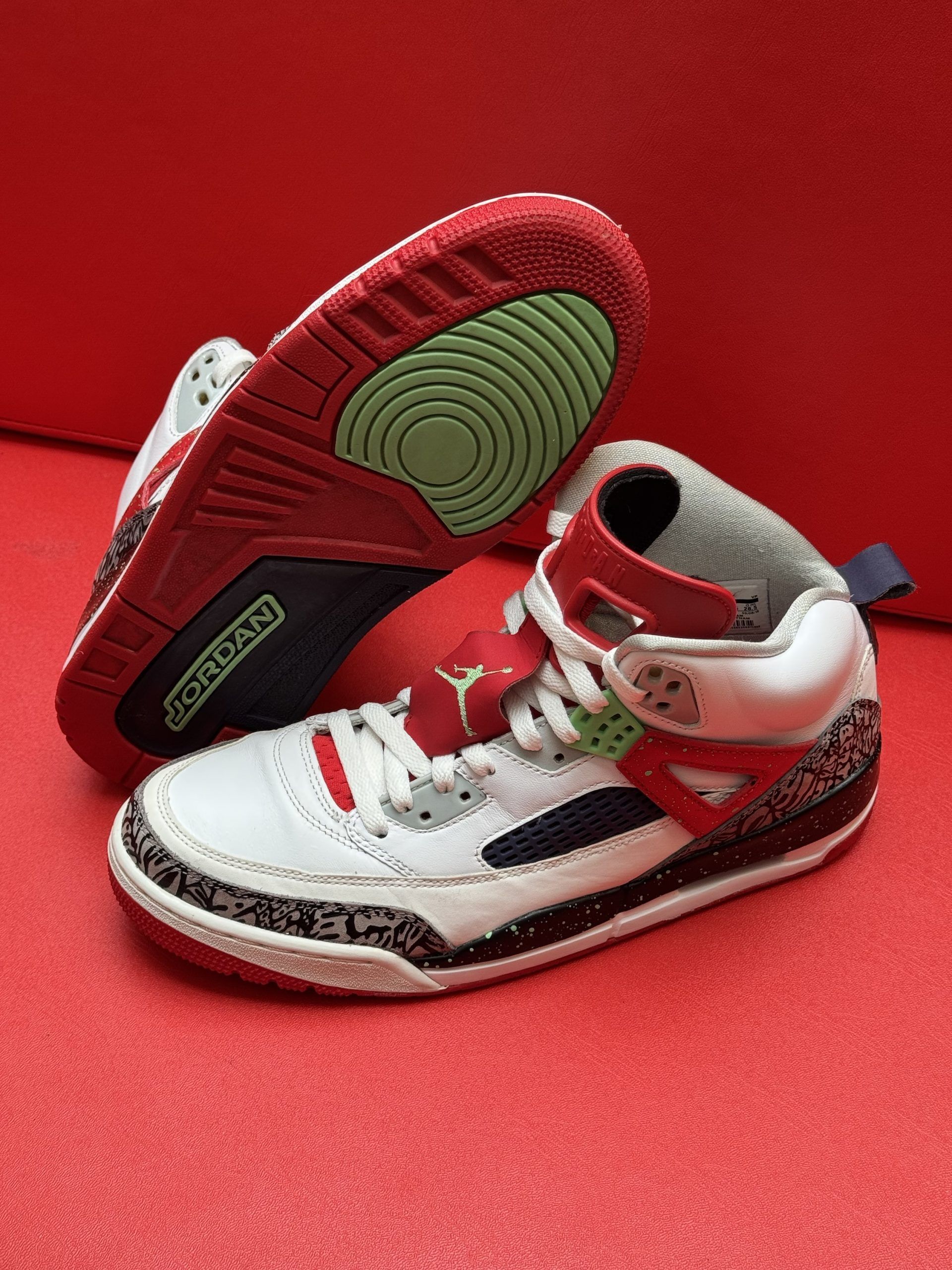 White, red, and blue Air Jordan sneakers on a red surface, showcasing the sole with green and red accents.