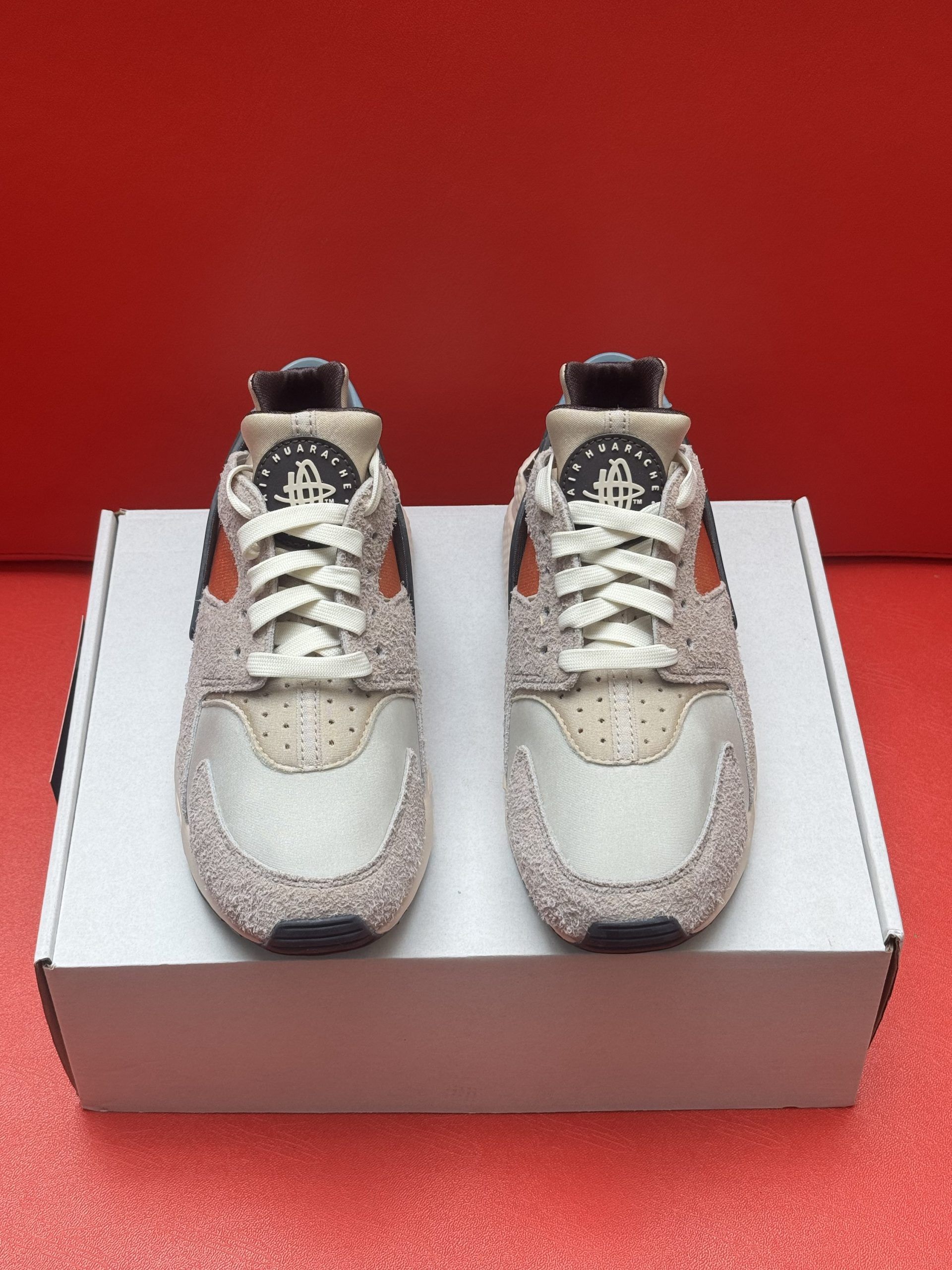 Pair of beige and orange Nike Huarache sneakers on a white box against a red background.