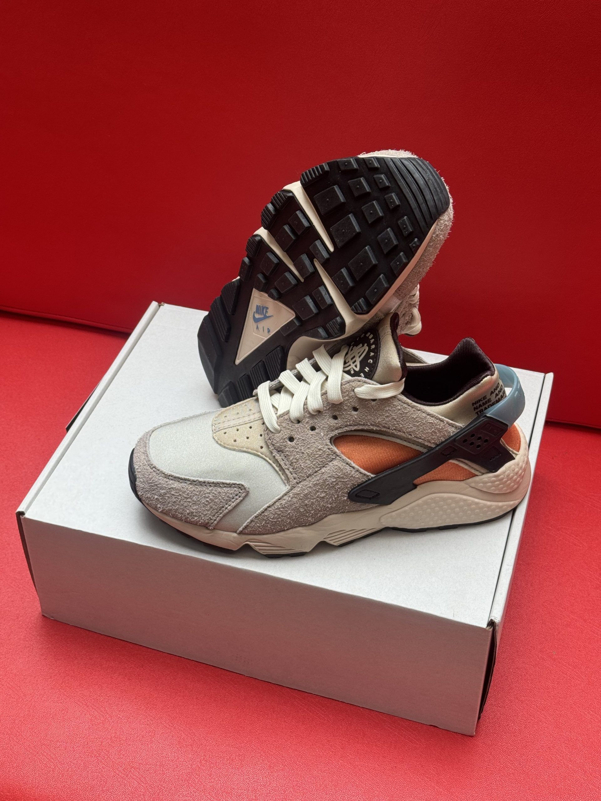 Pair of beige and orange Nike Huarache sneakers on a white box against a red background.