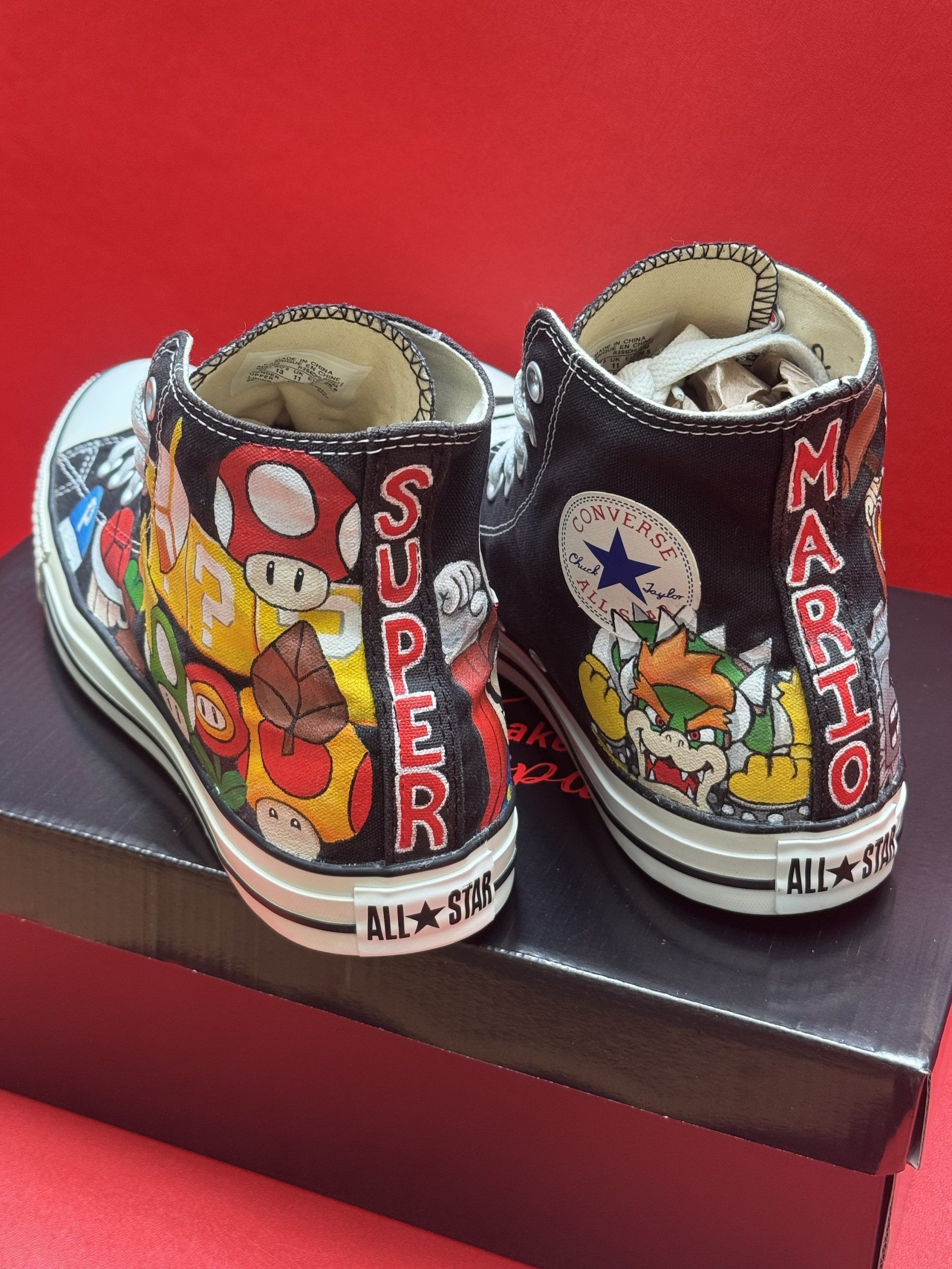 Black Converse high-tops painted with Super Mario characters, resting on a black box, against a red background.