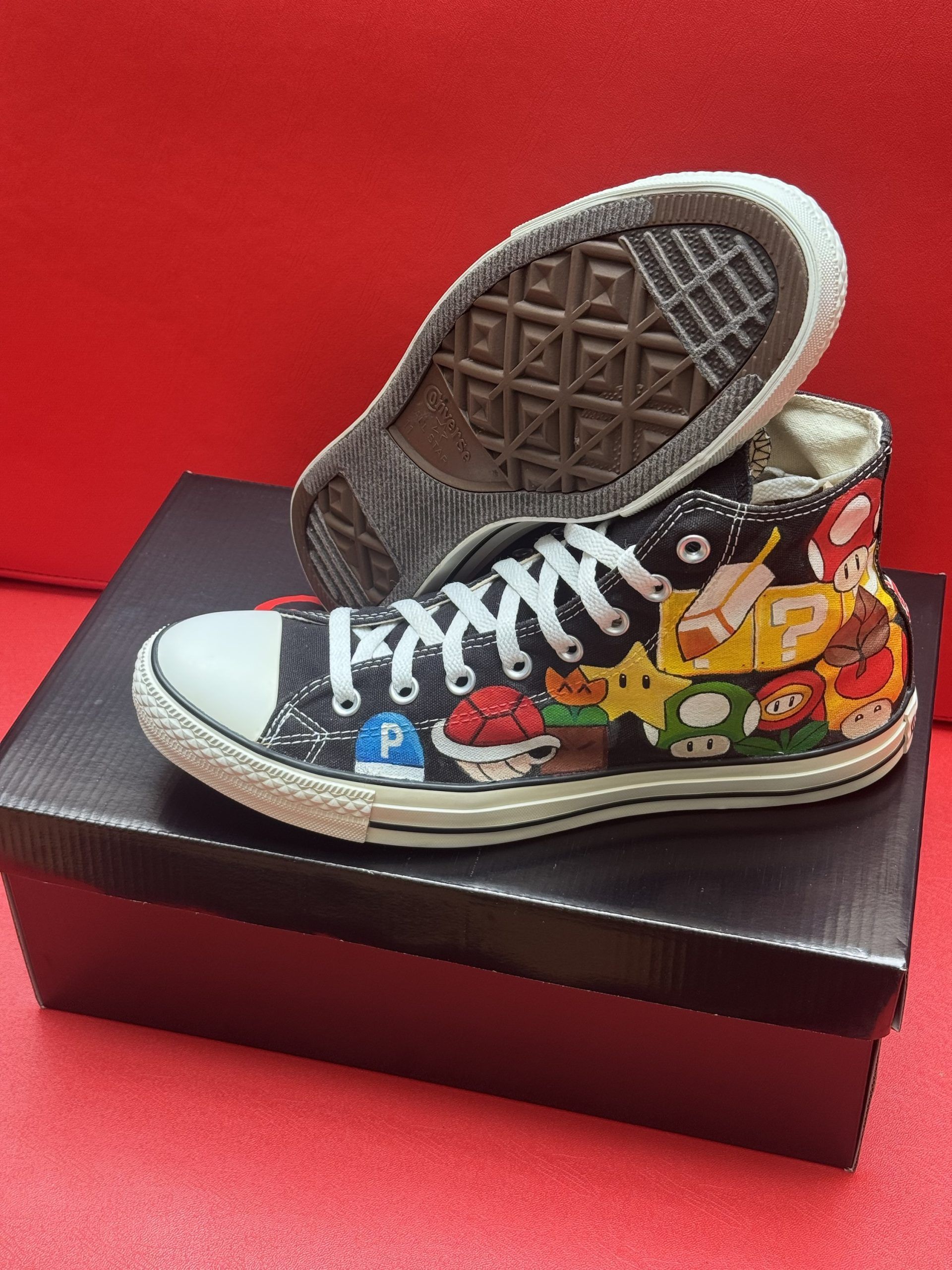 Black Converse high-tops painted with Super Mario characters, resting on a black box, against a red background.