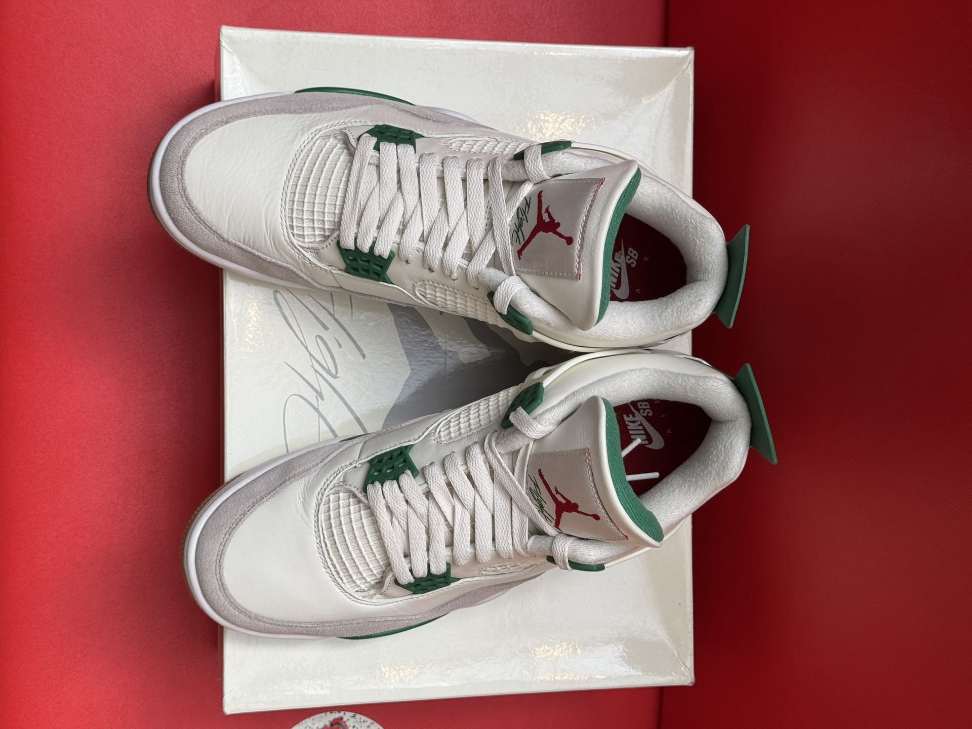White, green, and grey Air Jordan 4 sneakers on a box, red background. Gum soles.