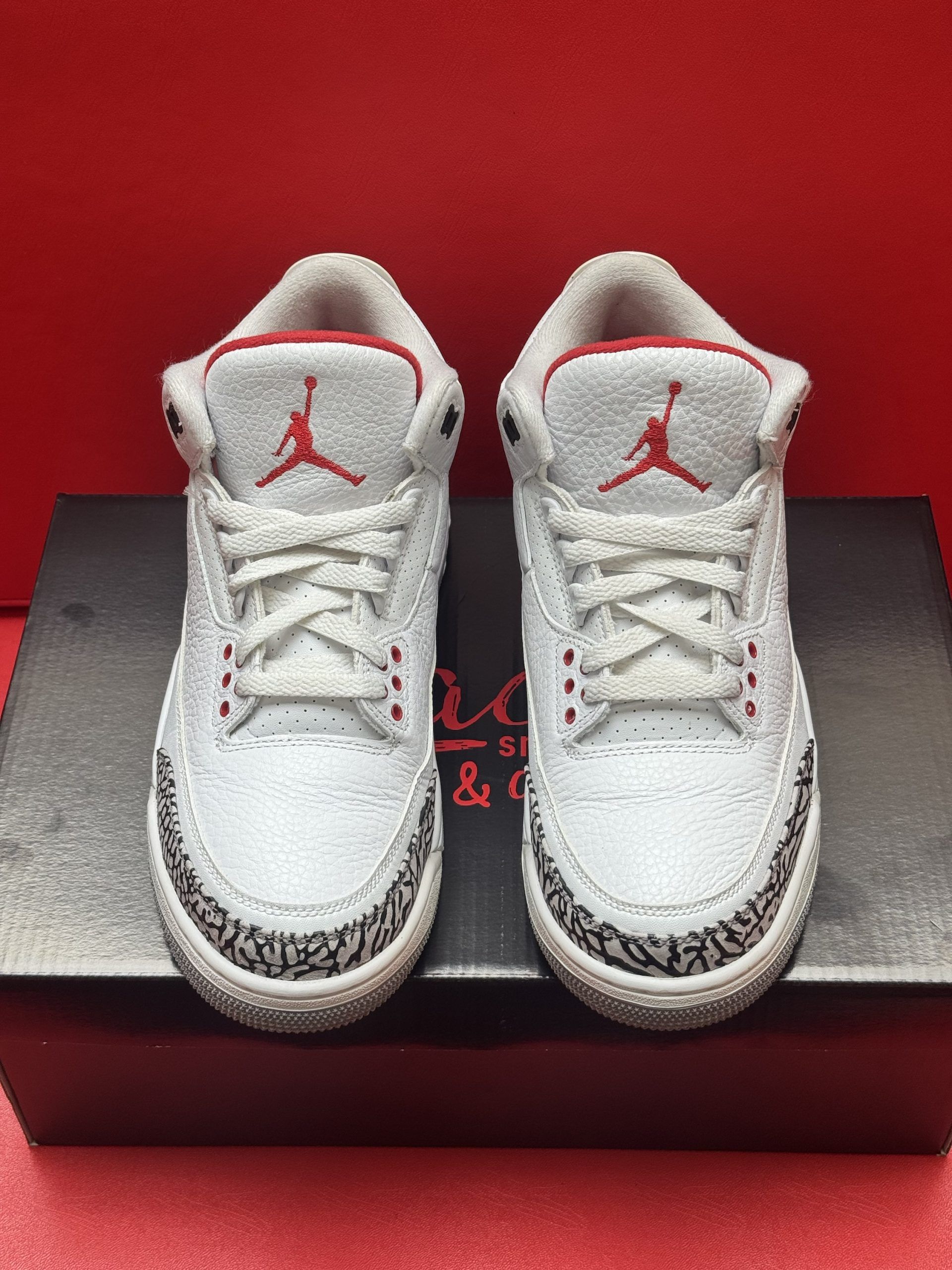 White and red Air Jordan 3 infant sneakers on a black shoebox against a red background.