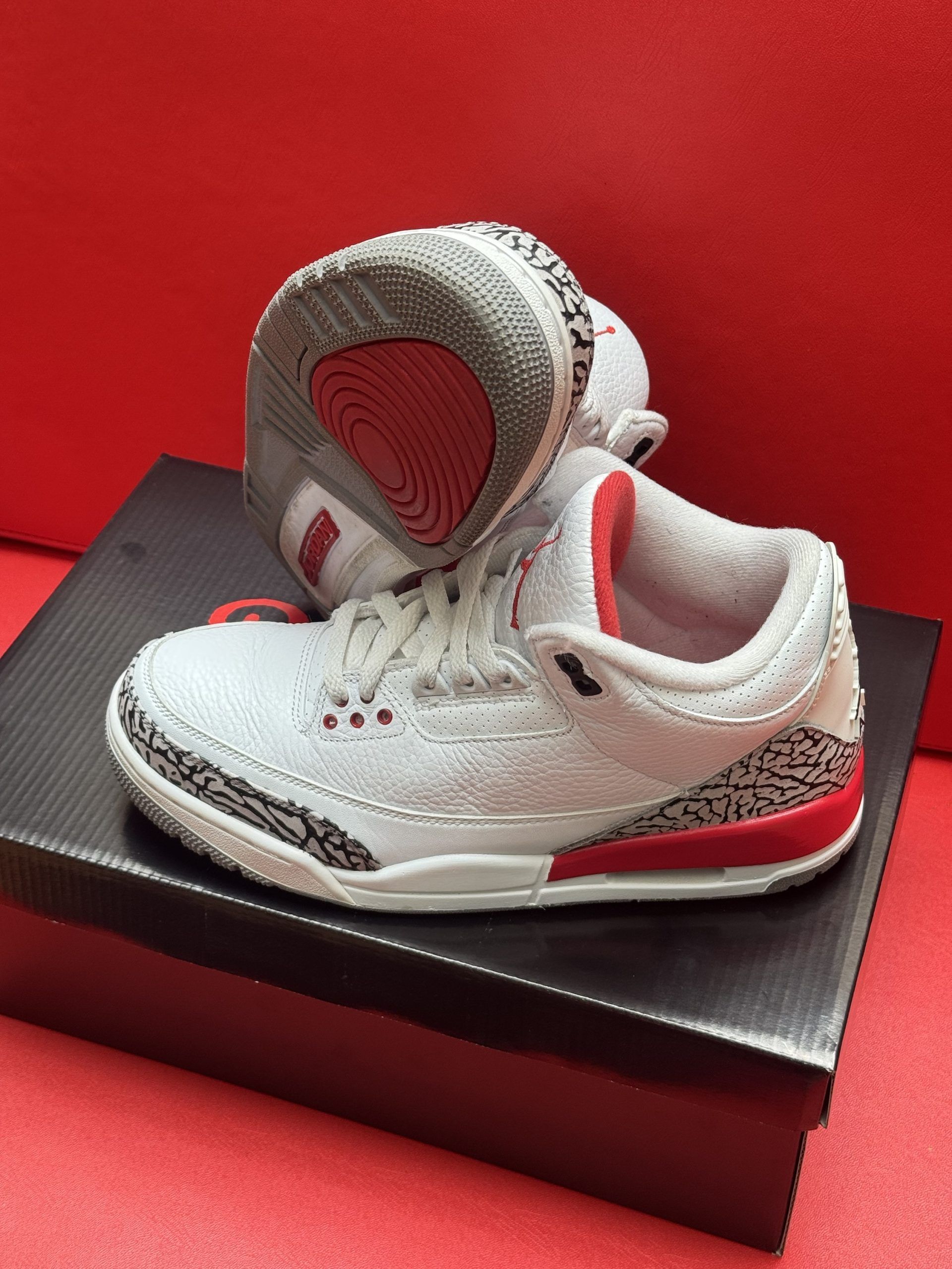 White and red Air Jordan 3 infant sneakers on a black shoebox against a red background.