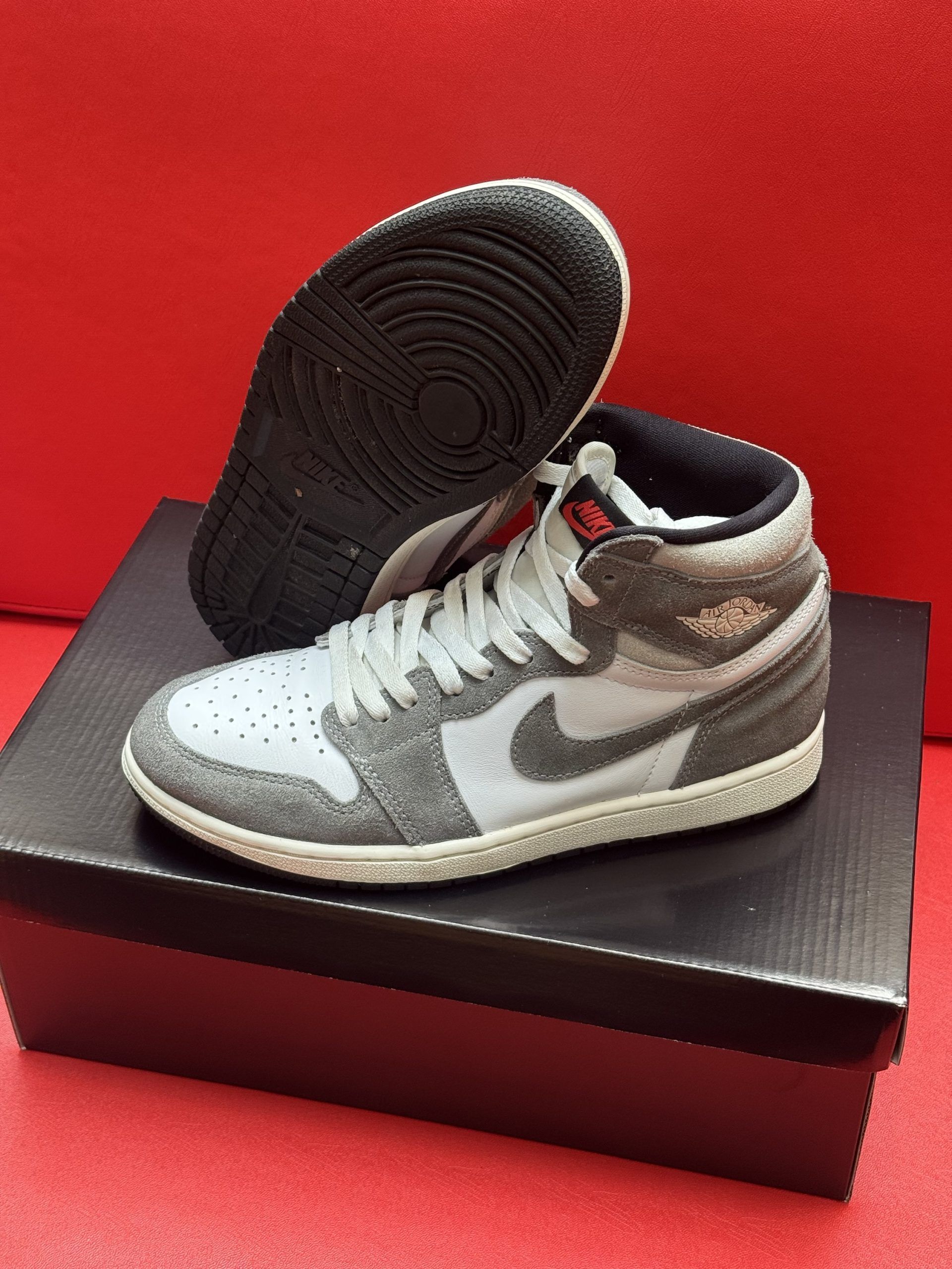 Gray and white Air Jordan 1 high-top sneakers on a black shoe box, set against a red background.
