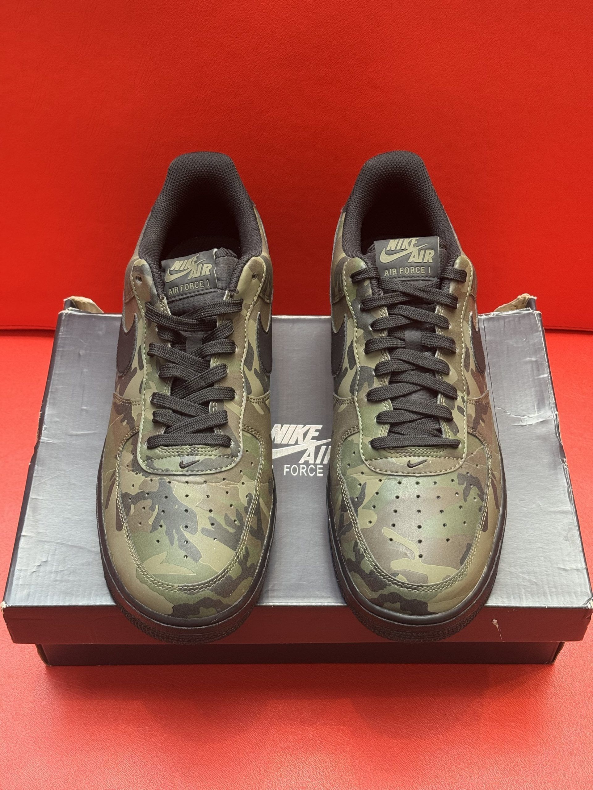 Olive green and black camouflage Nike Air Force 1 sneakers on a black shoe box, with one sole up.