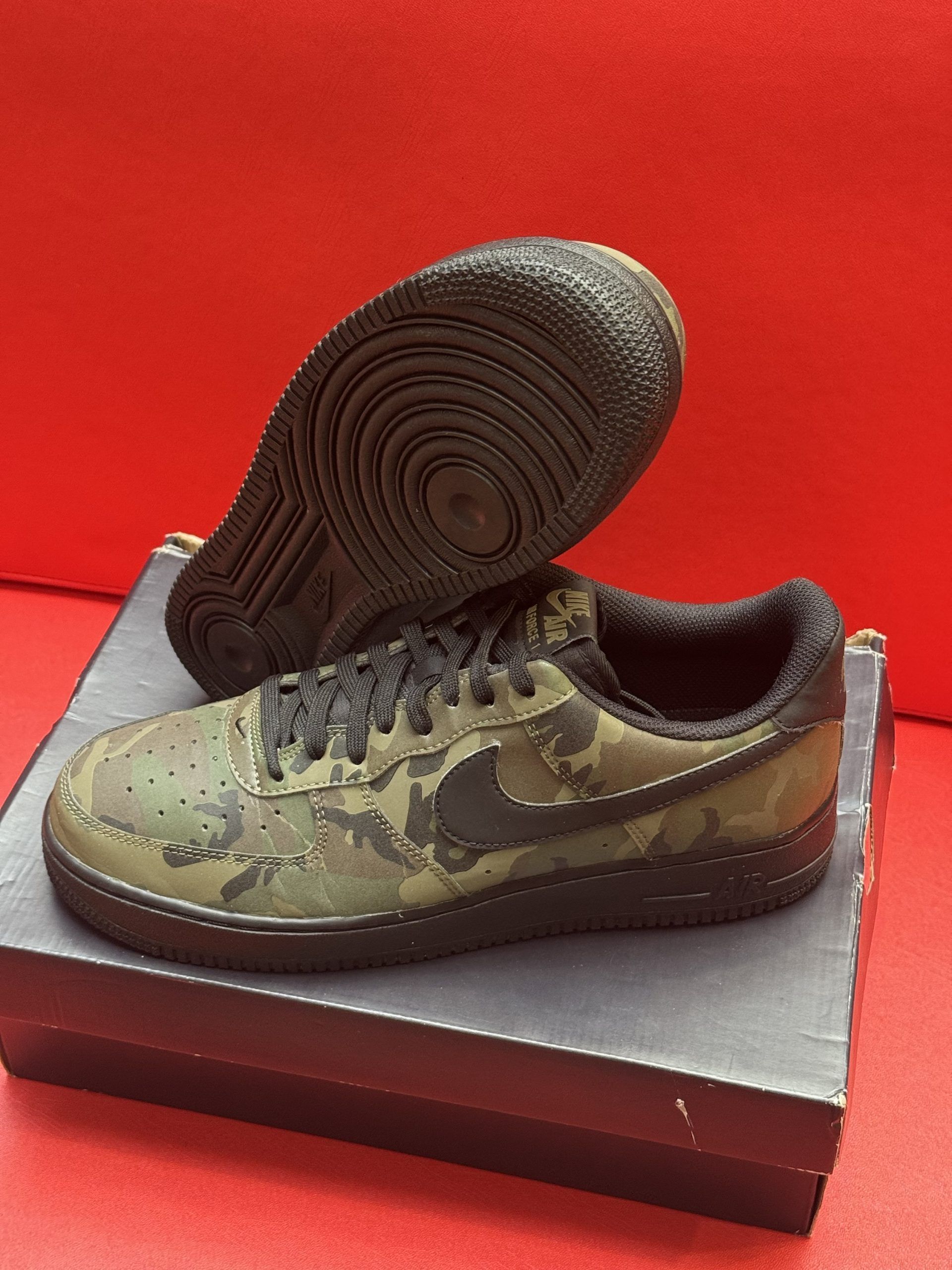 Olive green and black camouflage Nike Air Force 1 sneakers on a black shoe box, with one sole up.
