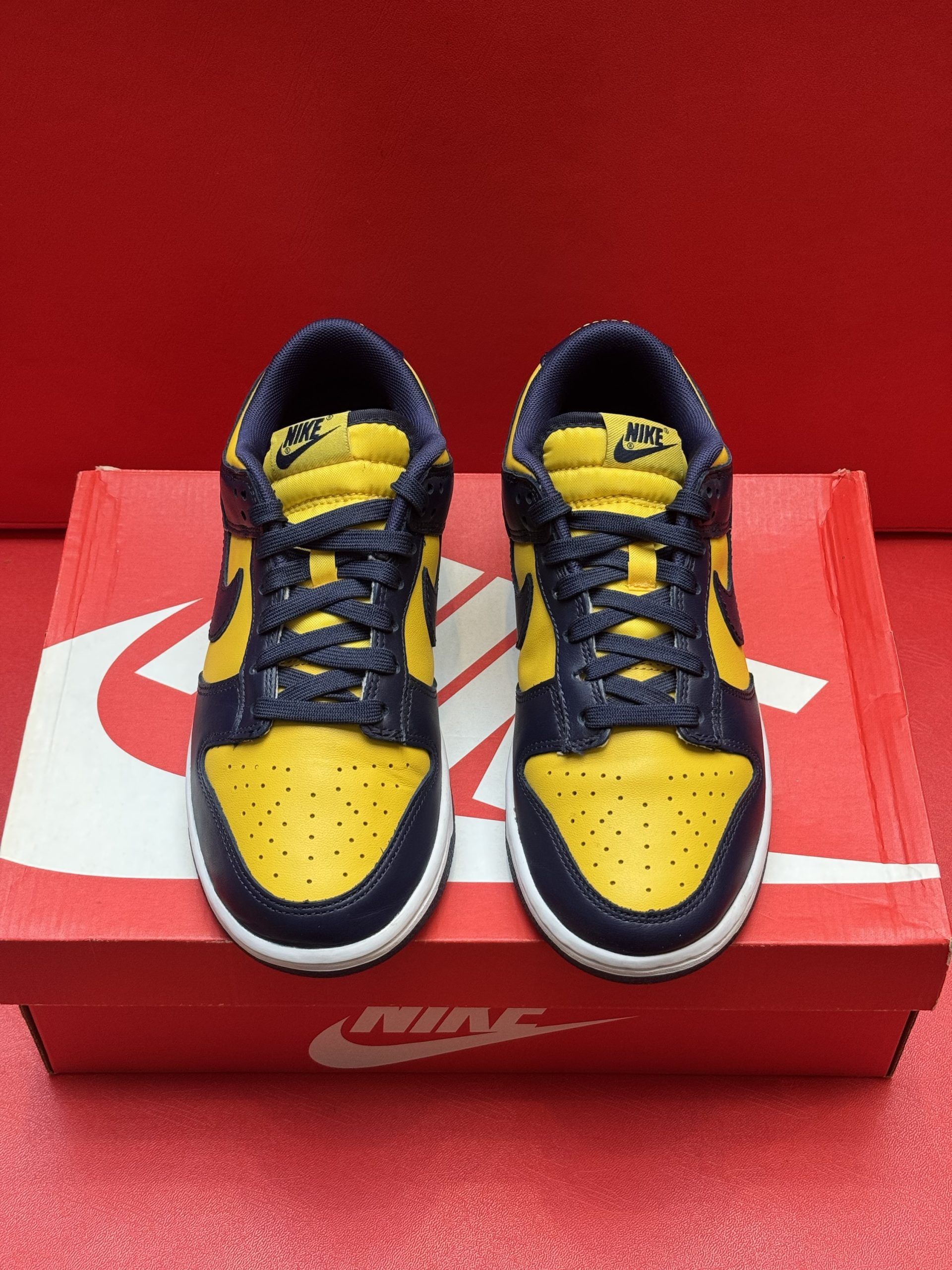 Yellow and navy Nike Dunk low sneakers on a red Nike box with a black sole against a red backdrop.