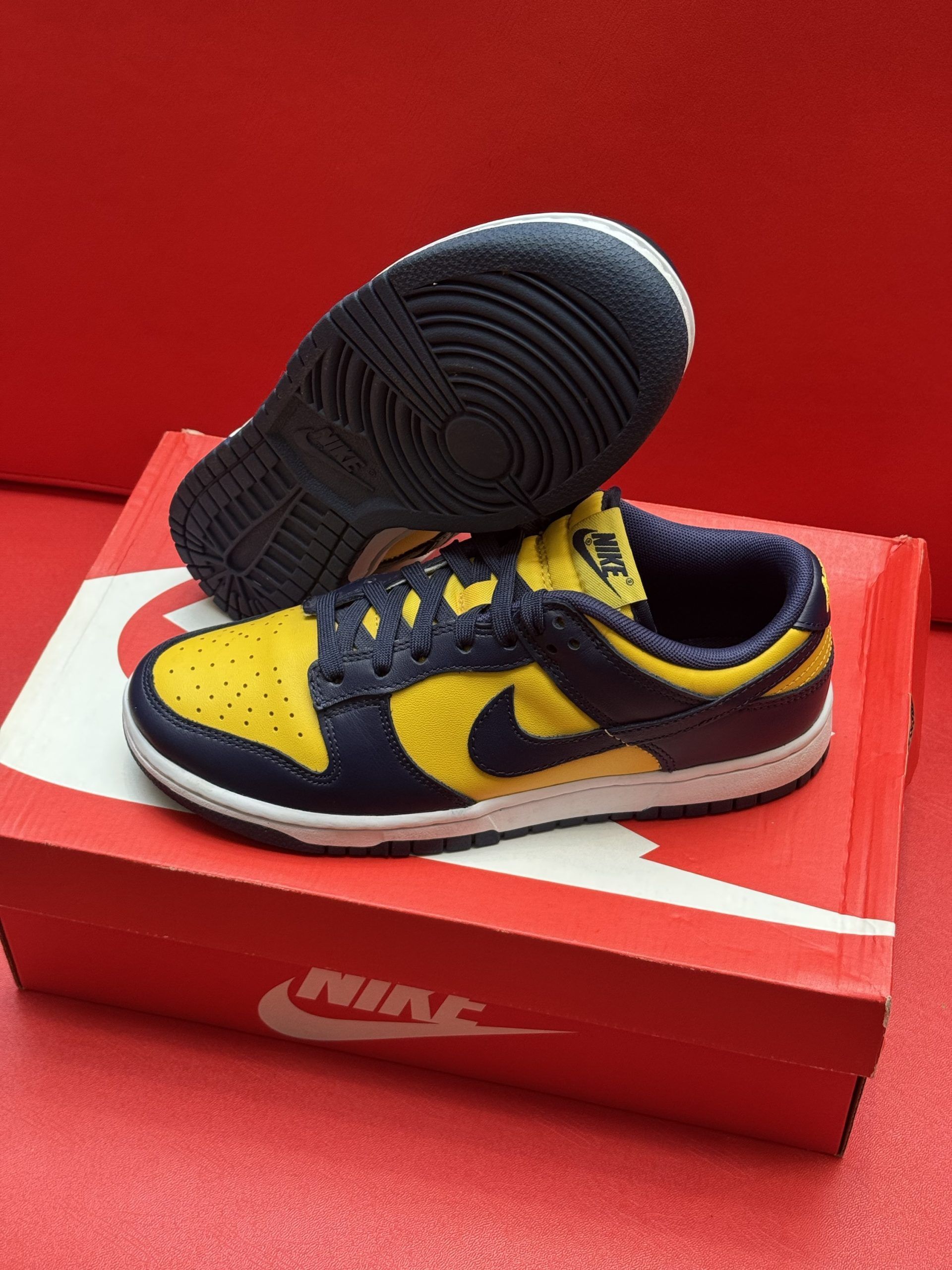 Yellow and navy Nike Dunk low sneakers on a red Nike box with a black sole against a red backdrop.