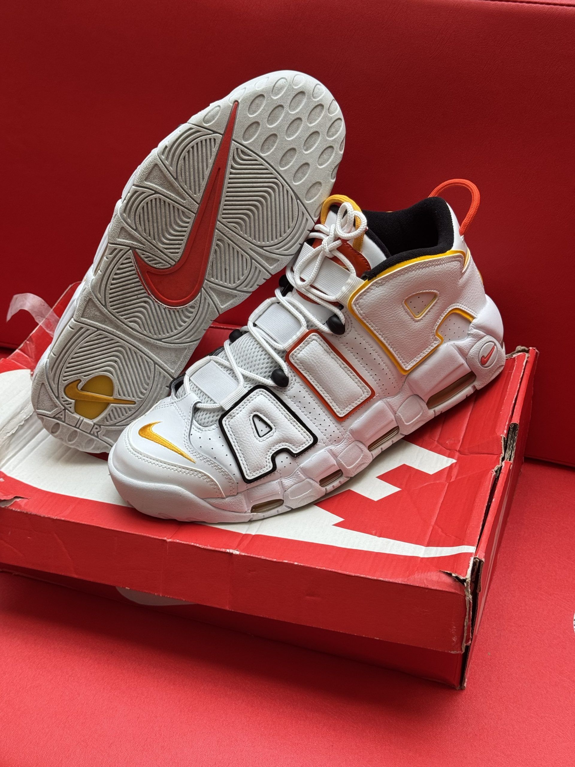 White Nike Air More Uptempo sneakers on a red box with red sole, orange and yellow accents.