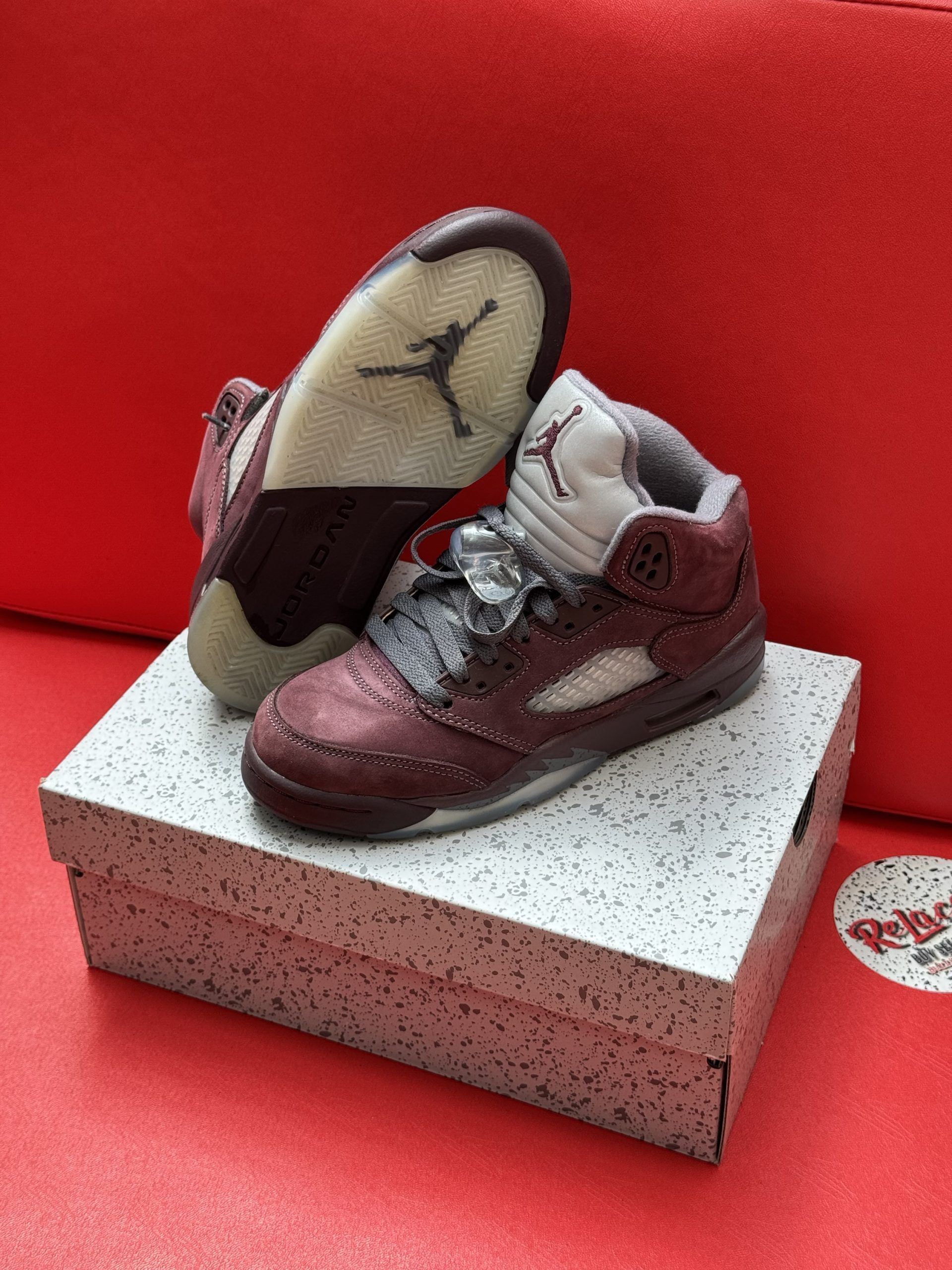 Air Jordan 5 sneakers, burgundy and gray, on a speckled box, set against a red background.