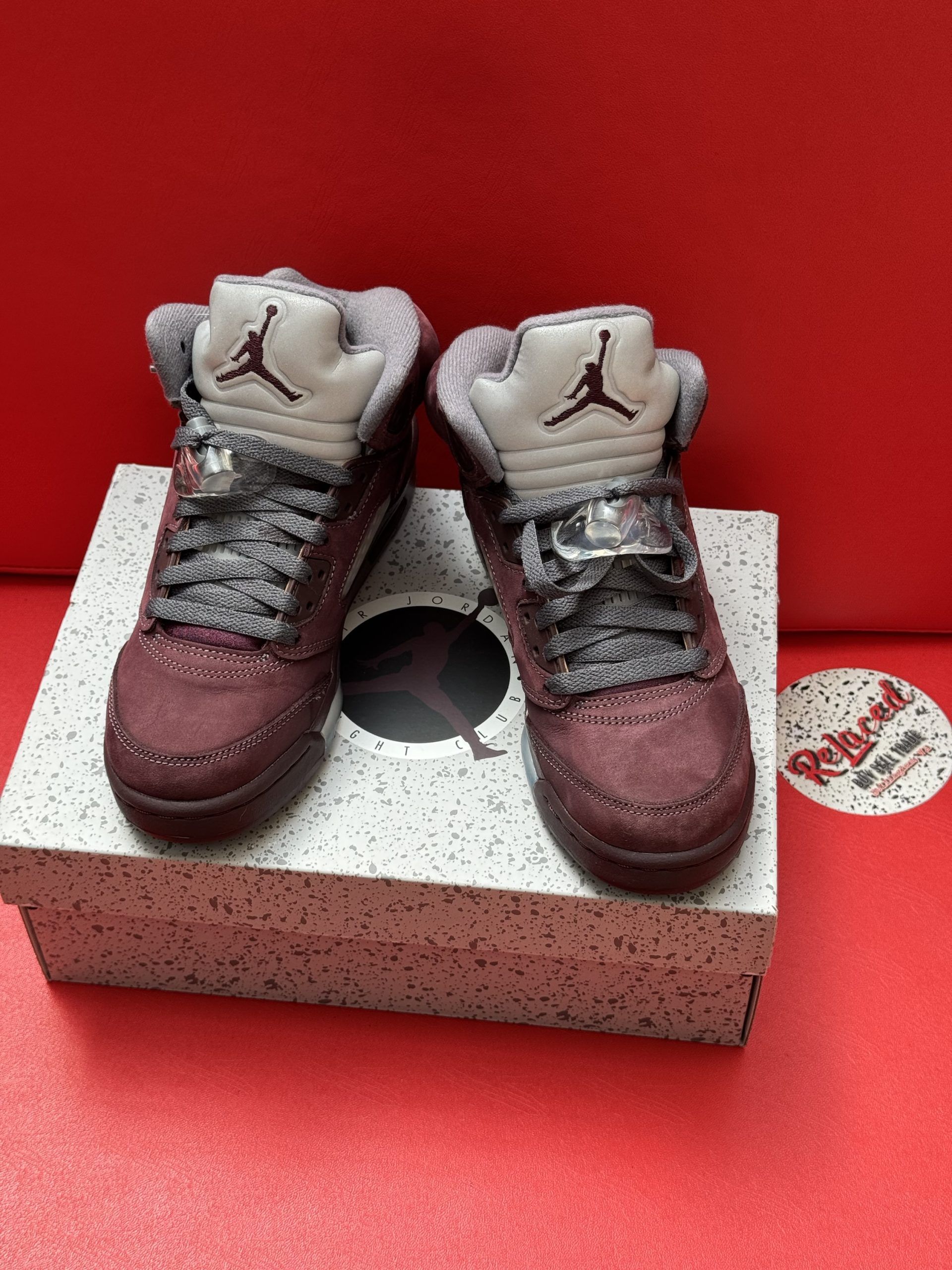 Air Jordan 5 sneakers, burgundy and gray, on a speckled box, set against a red background.
