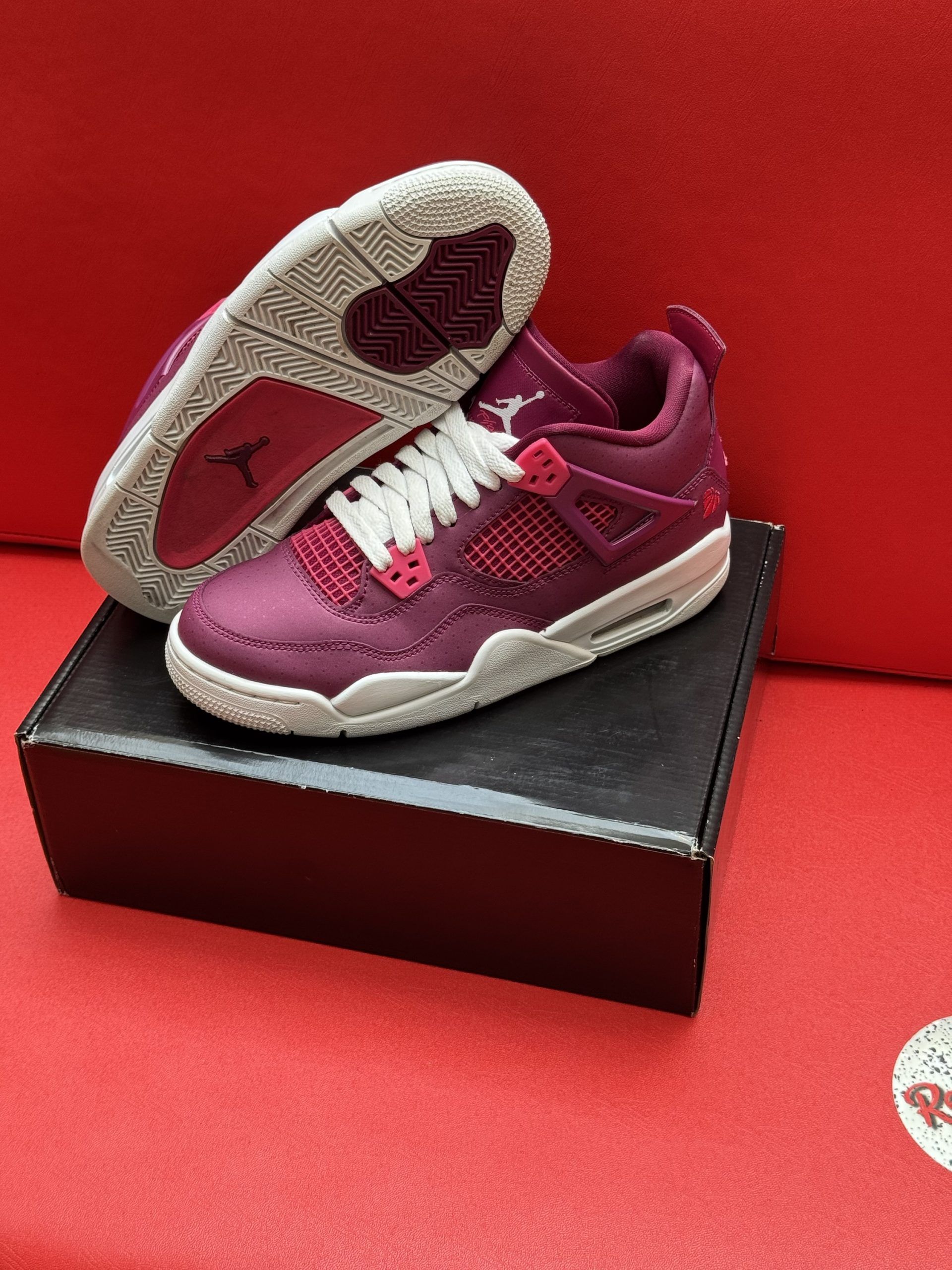 Pair of burgundy and white Air Jordan 4 sneakers on a black box, set against a red background.
