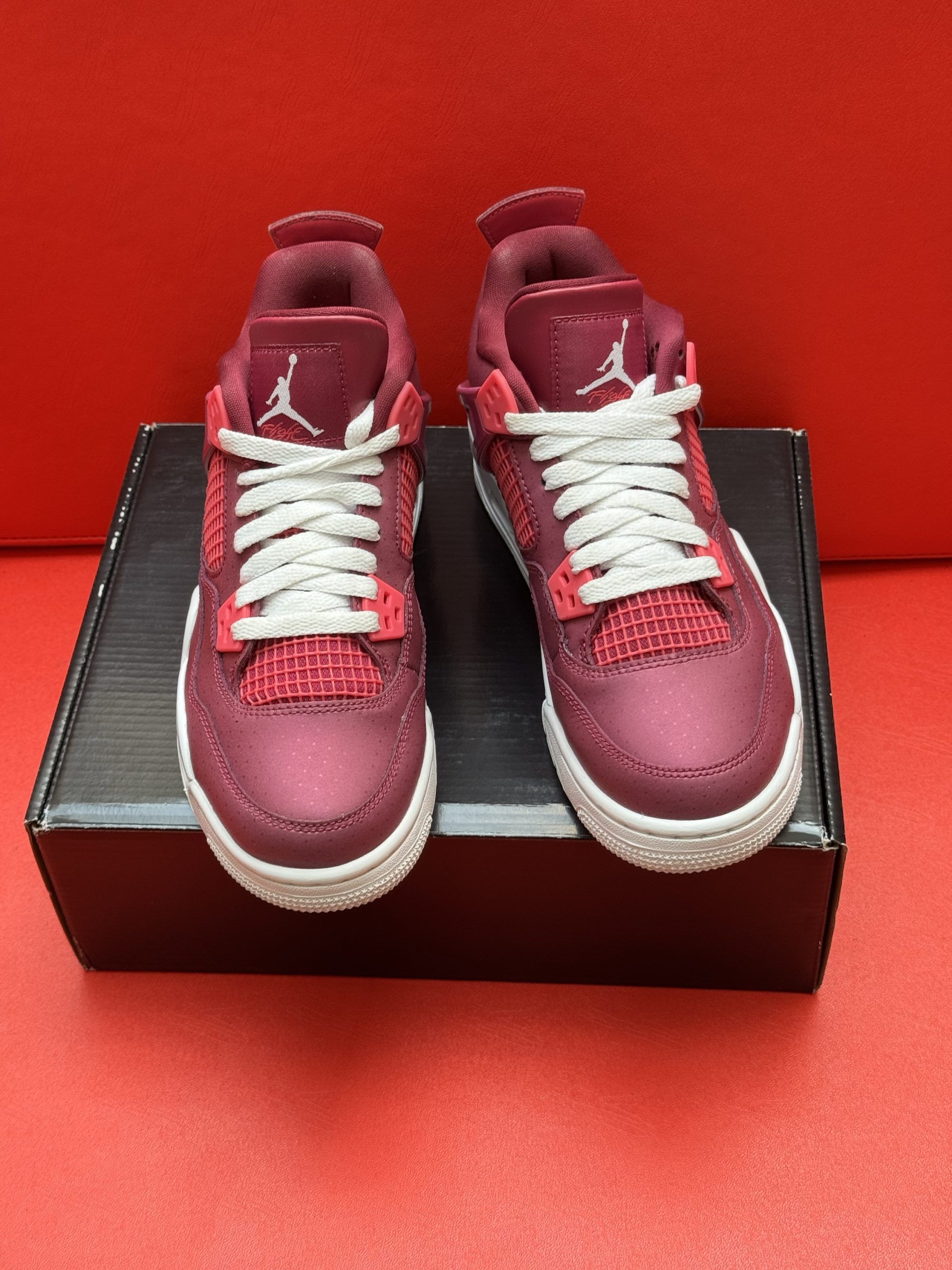 Pair of burgundy and white Air Jordan 4 sneakers on a black box, set against a red background.