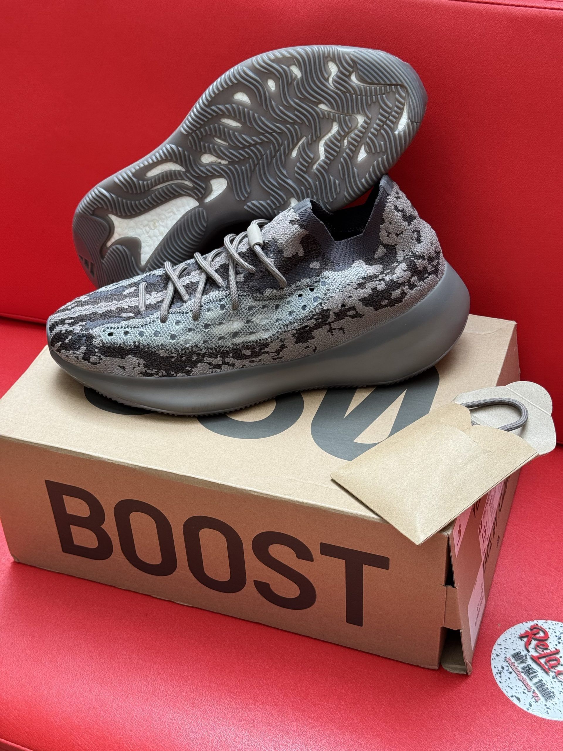 Brown and gray Adidas Yeezy sneakers on a cardboard box, with the sole visible. Red surface.