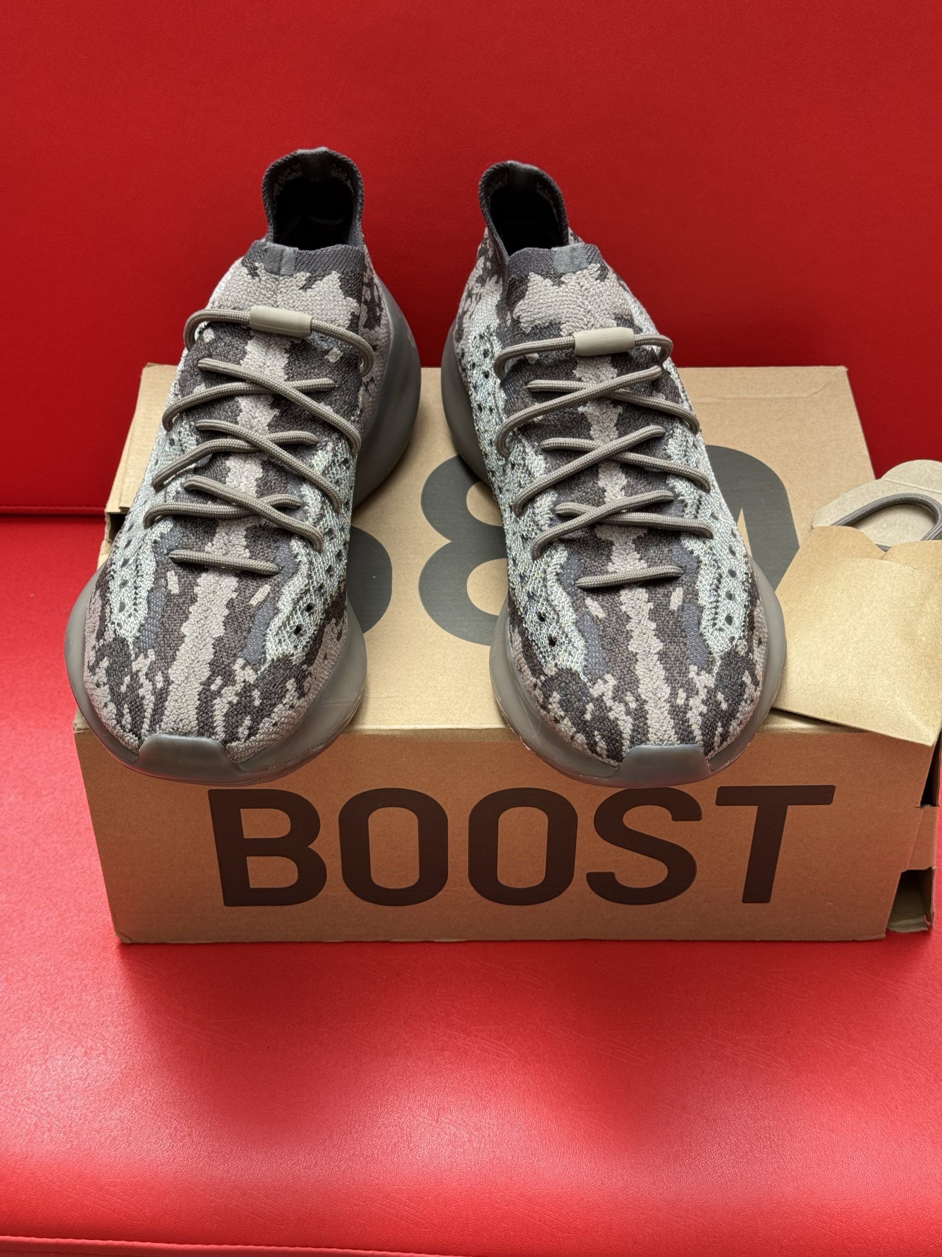 Brown and gray Adidas Yeezy sneakers on a cardboard box, with the sole visible. Red surface.
