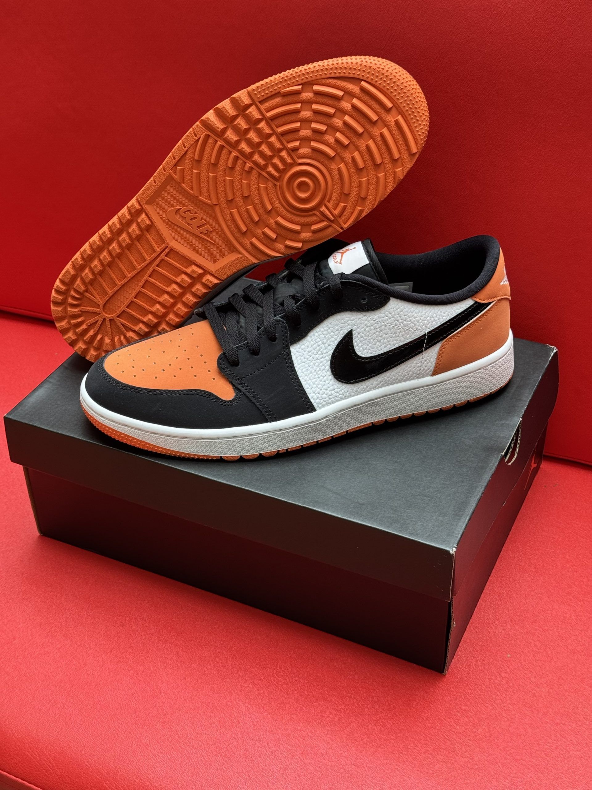 Orange, black, and white Air Jordan 1 Low sneaker displayed with the sole above the shoe on a box, red backdrop.
