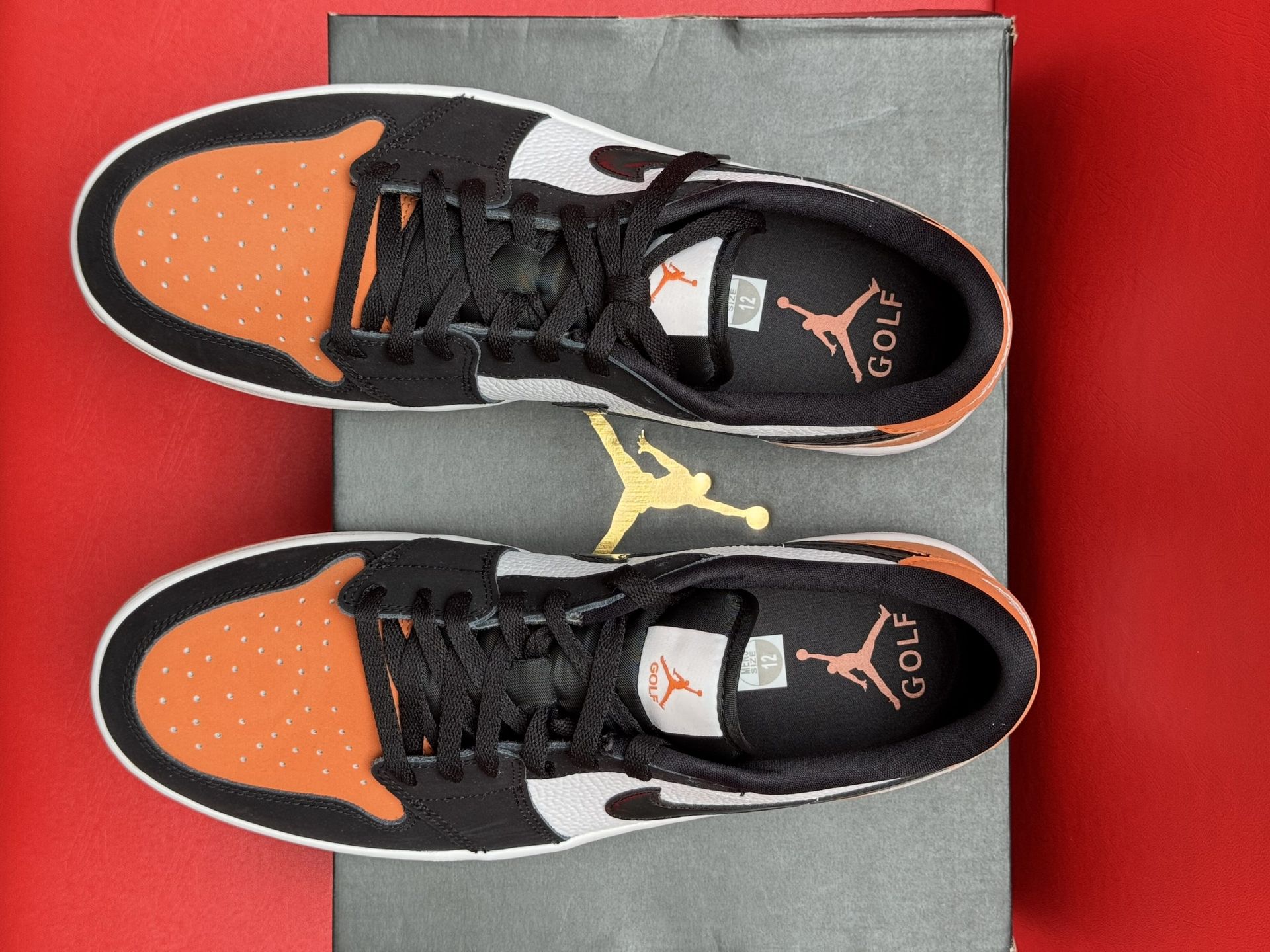 Orange, black, and white Air Jordan 1 Low sneaker displayed with the sole above the shoe on a box, red backdrop.