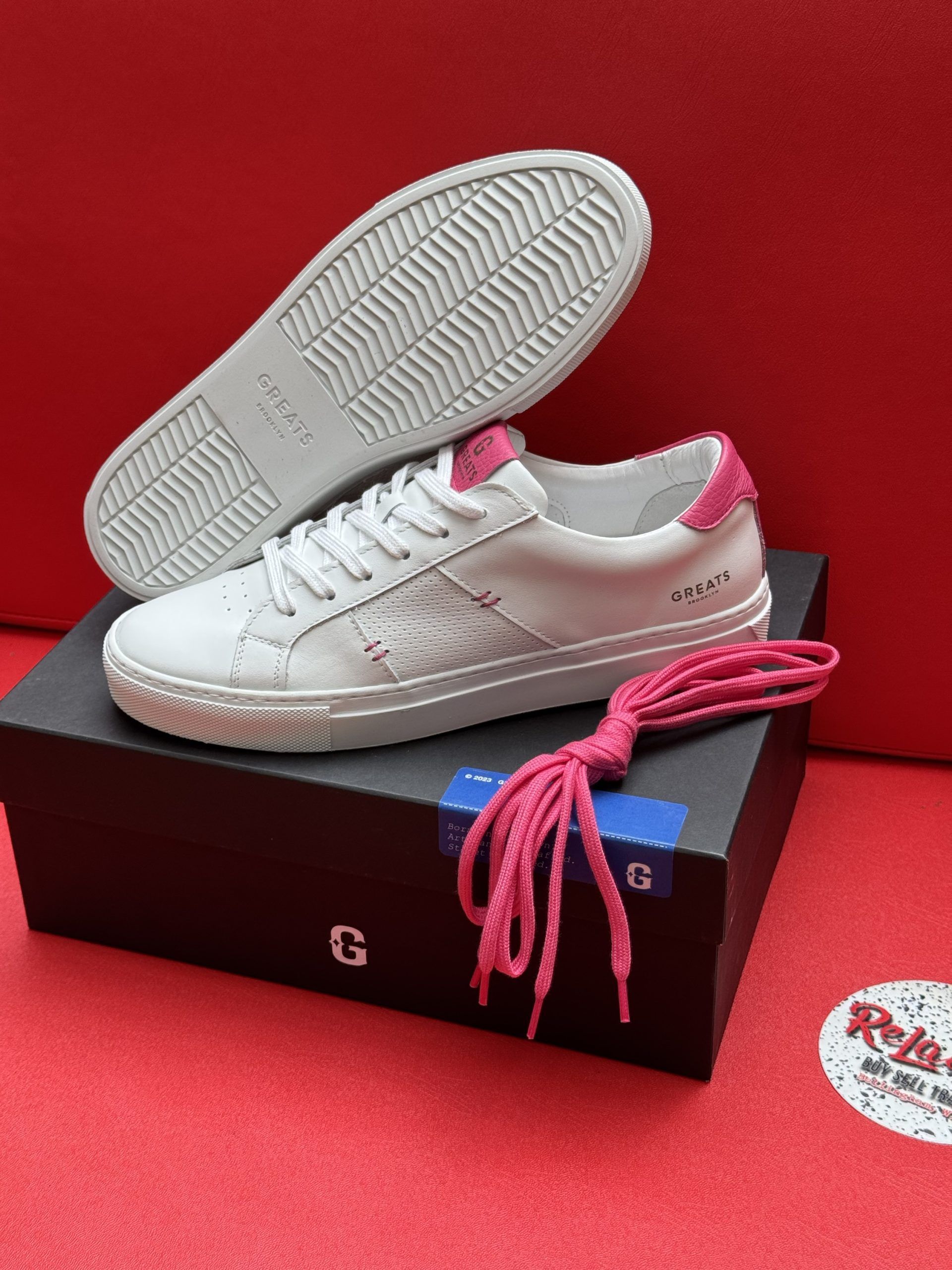White leather sneakers with pink accents, laces, and box on red surface.