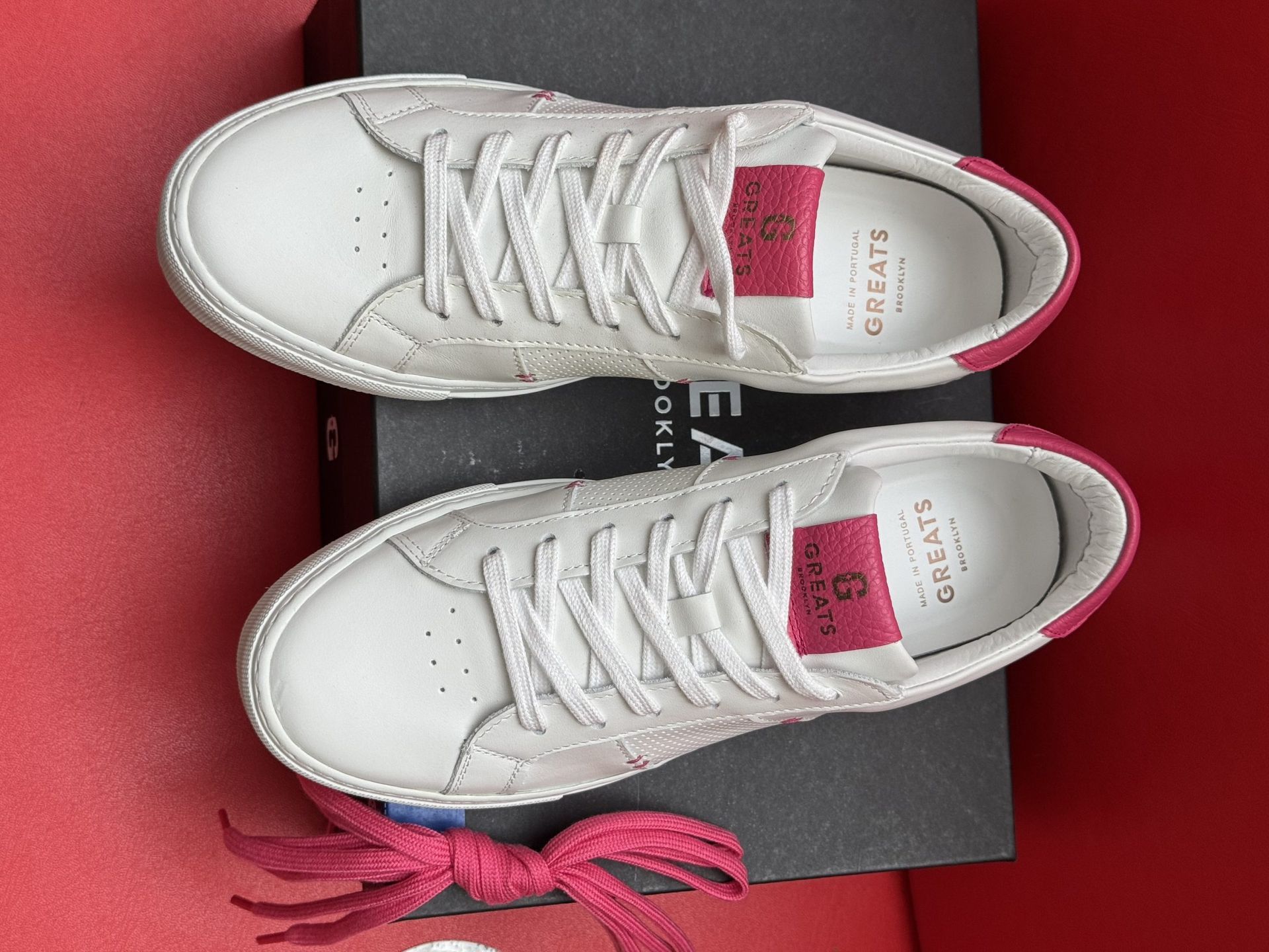 White leather sneakers with pink accents, laces, and box on red surface.
