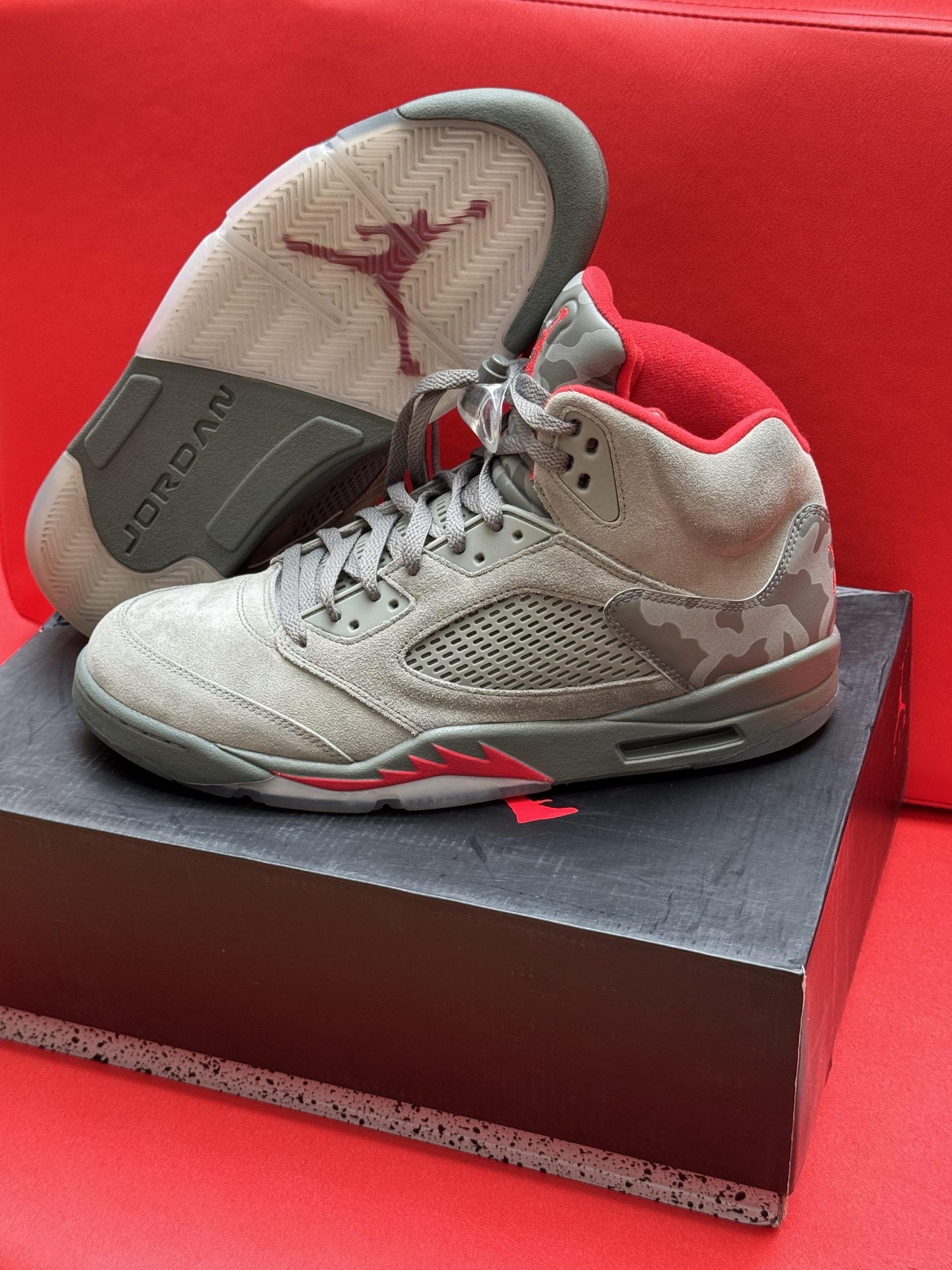 Gray and red Air Jordan 5 sneakers with a camouflage pattern, on a box, red background.