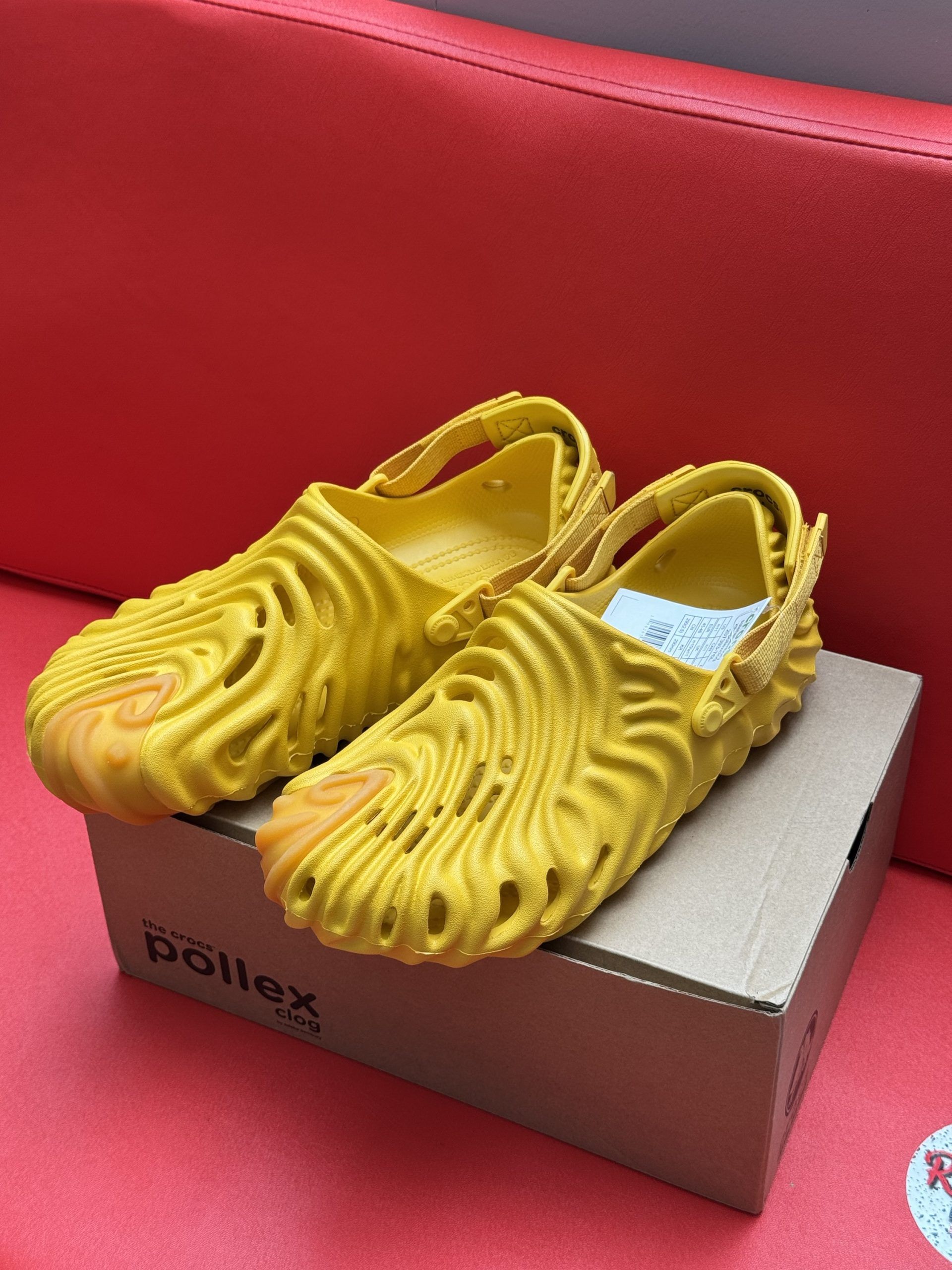 Yellow Pollex clogs with wavy design on a cardboard box, set against a red background.