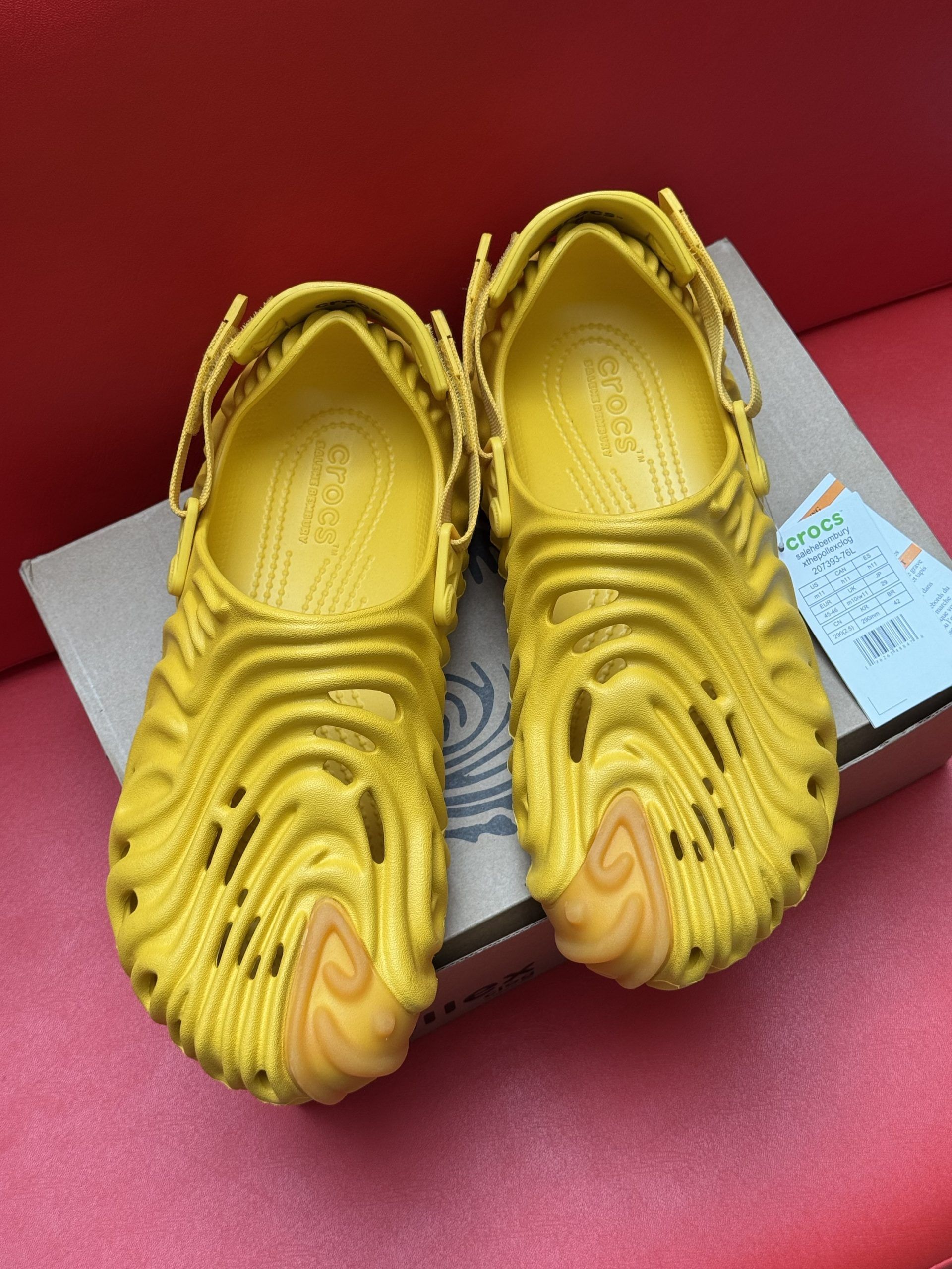 Yellow Pollex clogs with wavy design on a cardboard box, set against a red background.