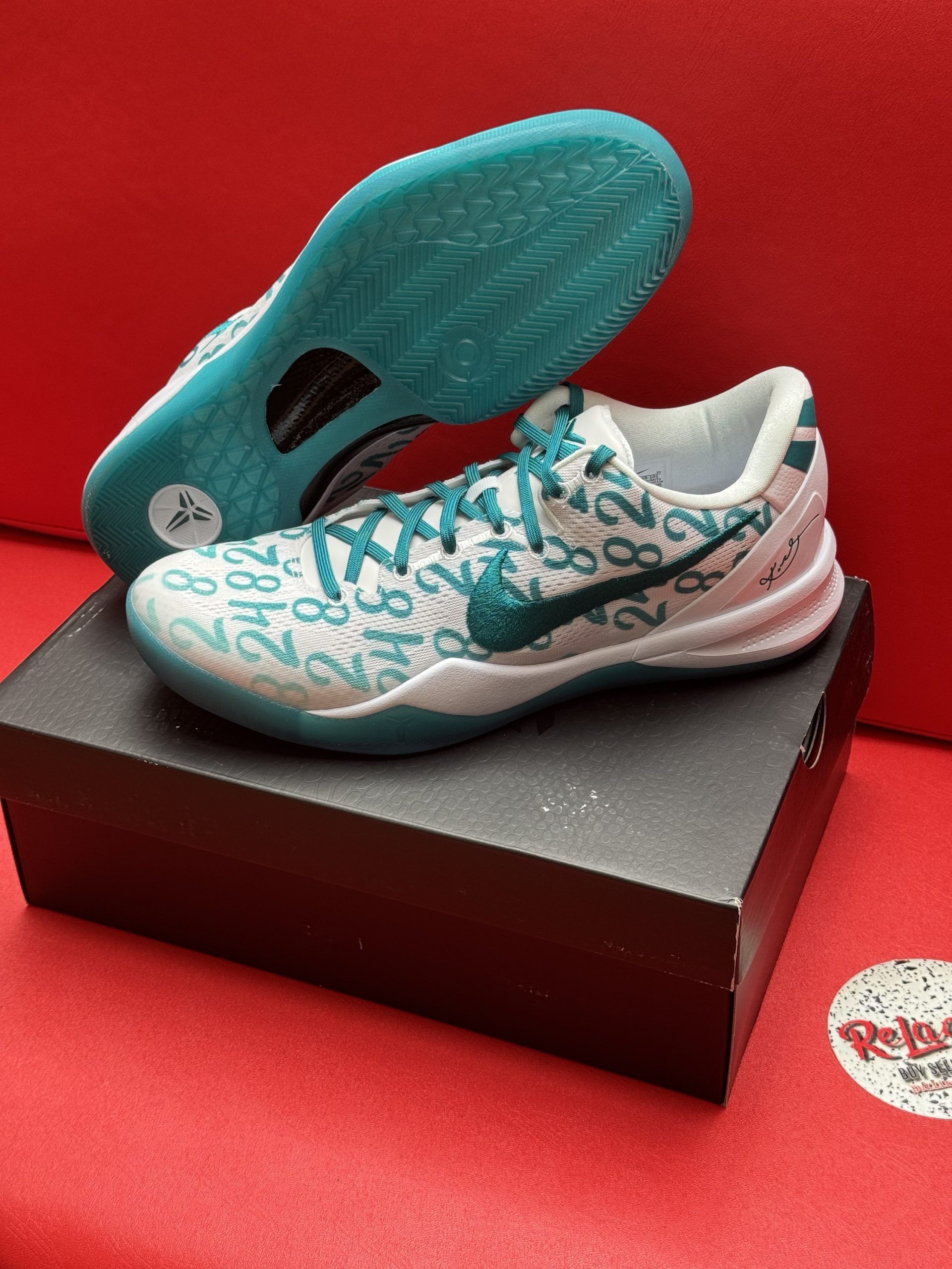 Nike Kobe Mamba Focus basketball sneakers on a box, white with teal accents and a teal sole, set against a red background.