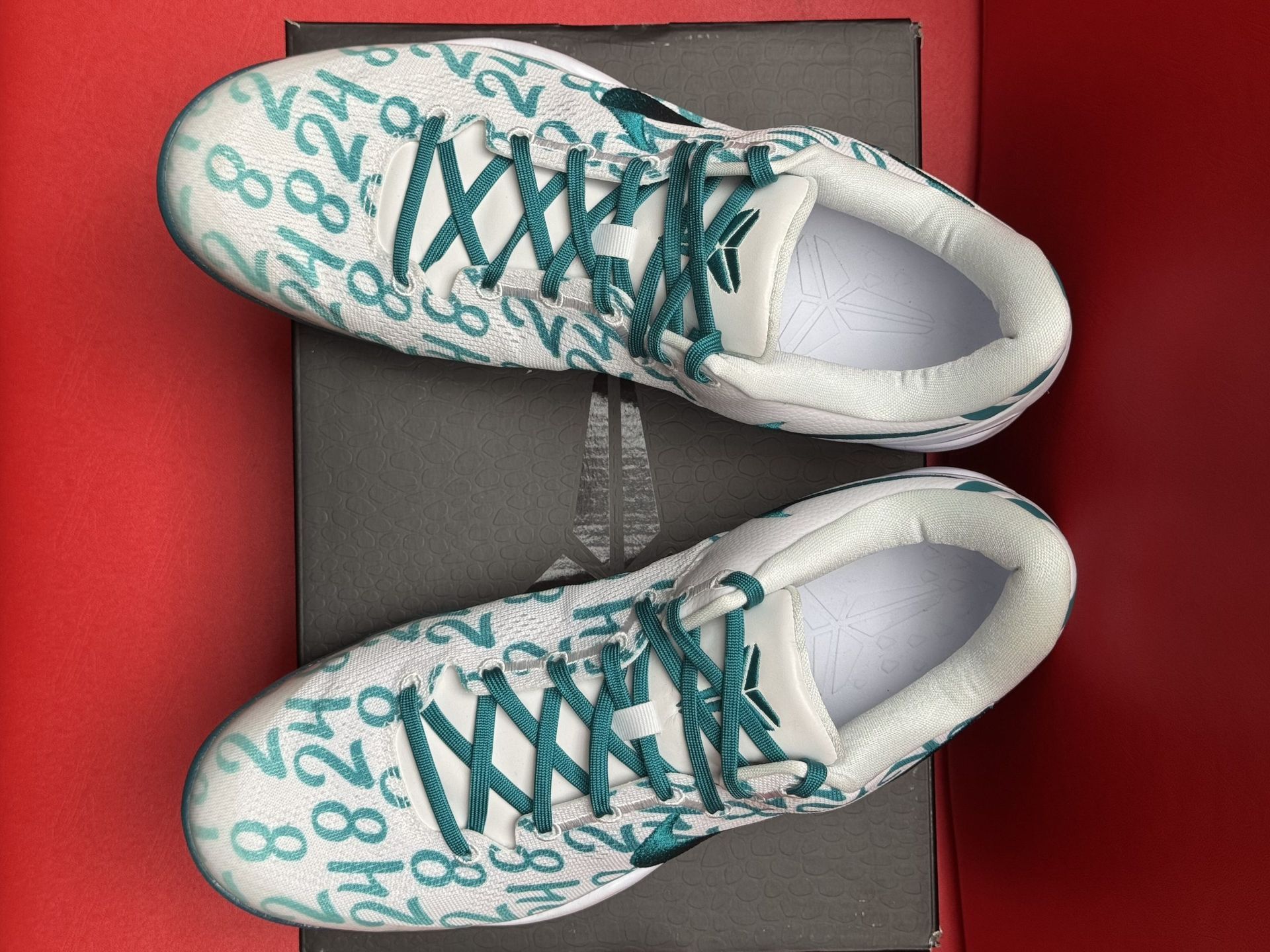 Nike Kobe Mamba Focus basketball sneakers on a box, white with teal accents and a teal sole, set against a red background.