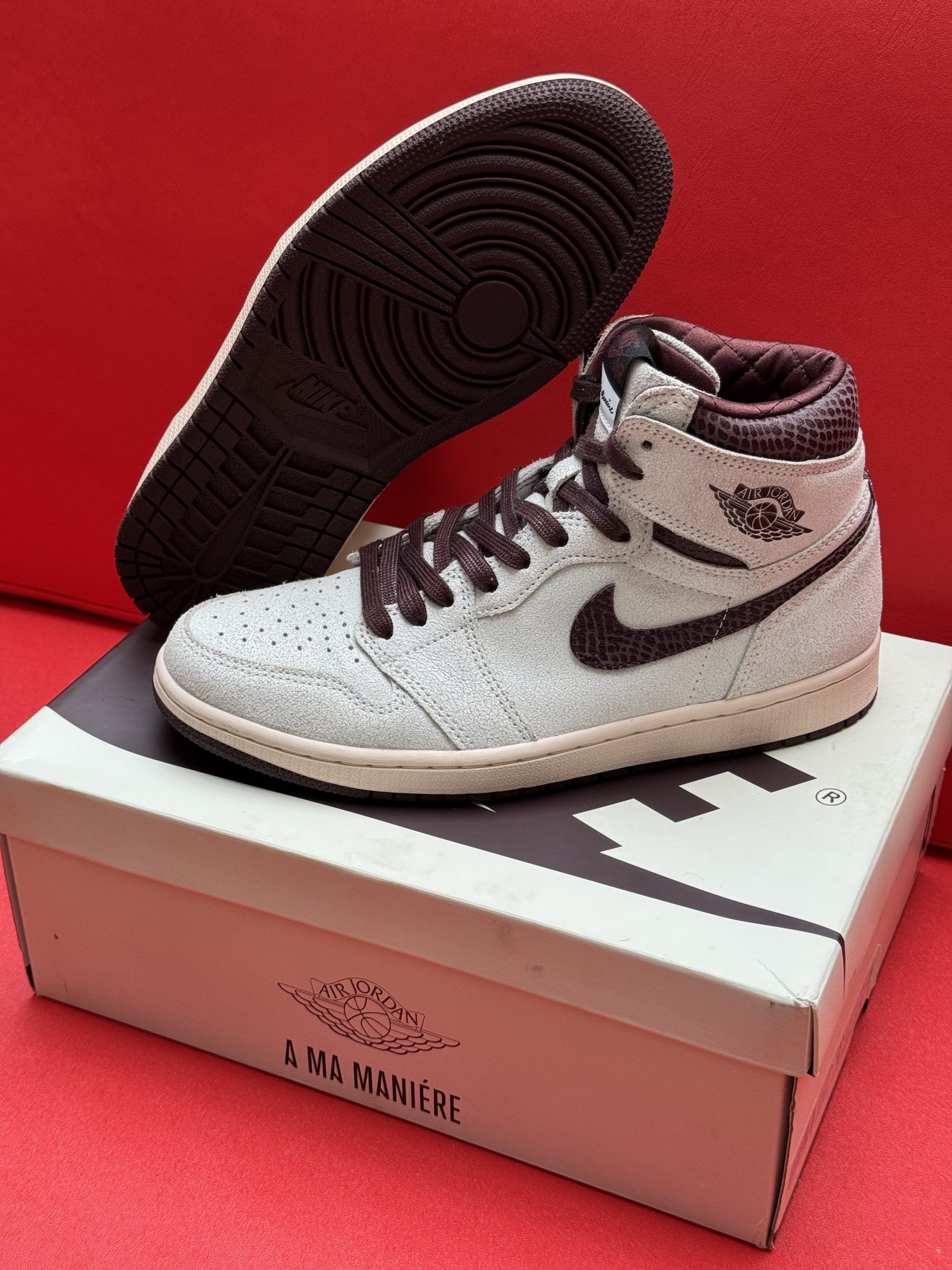 Air Jordan 1 high-top sneaker in white and dark red, on top of a box, with a red background.
