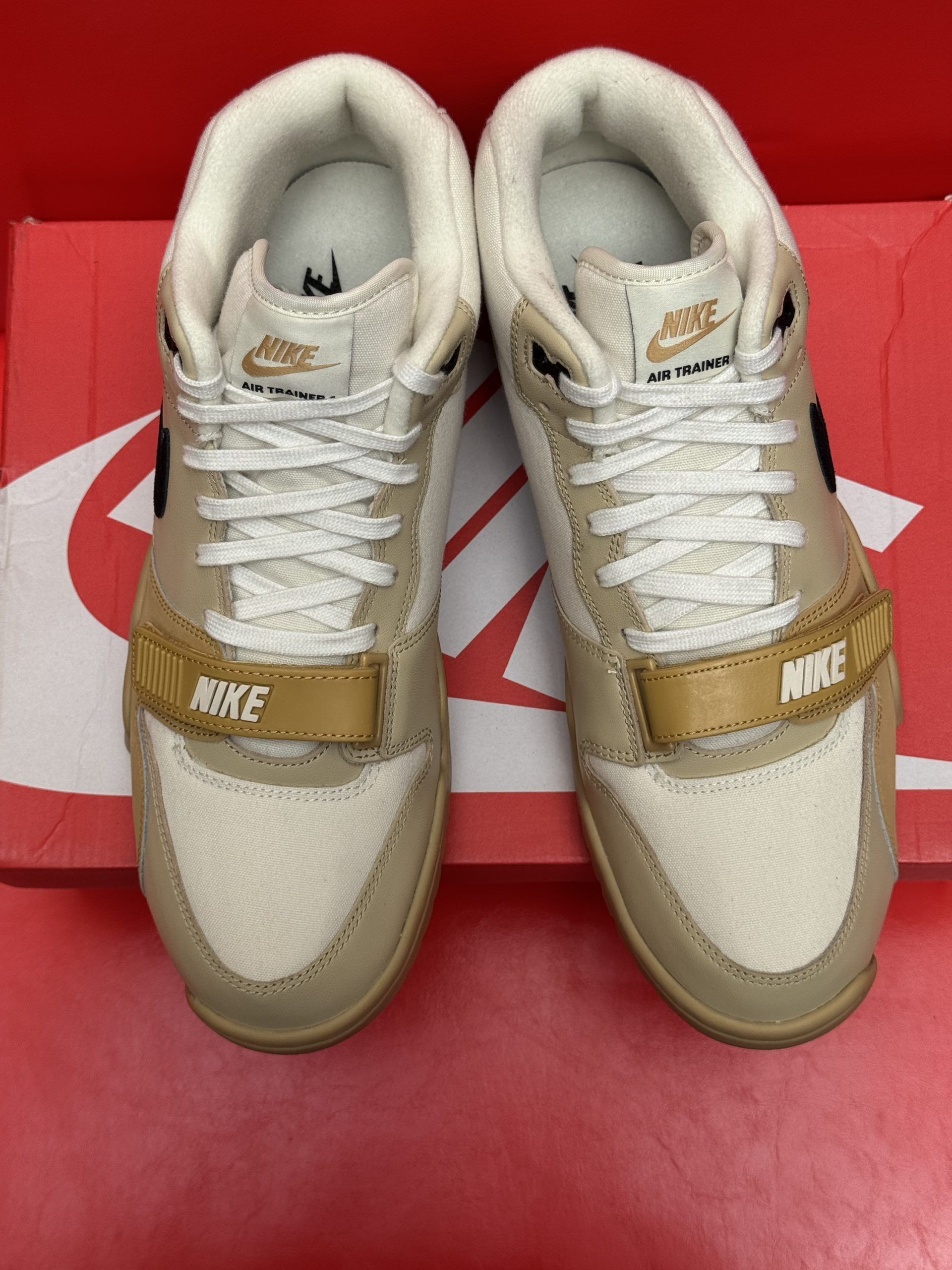 Beige and black Nike Air Trainer sneakers on a red Nike box with a beige sole, against a red surface.