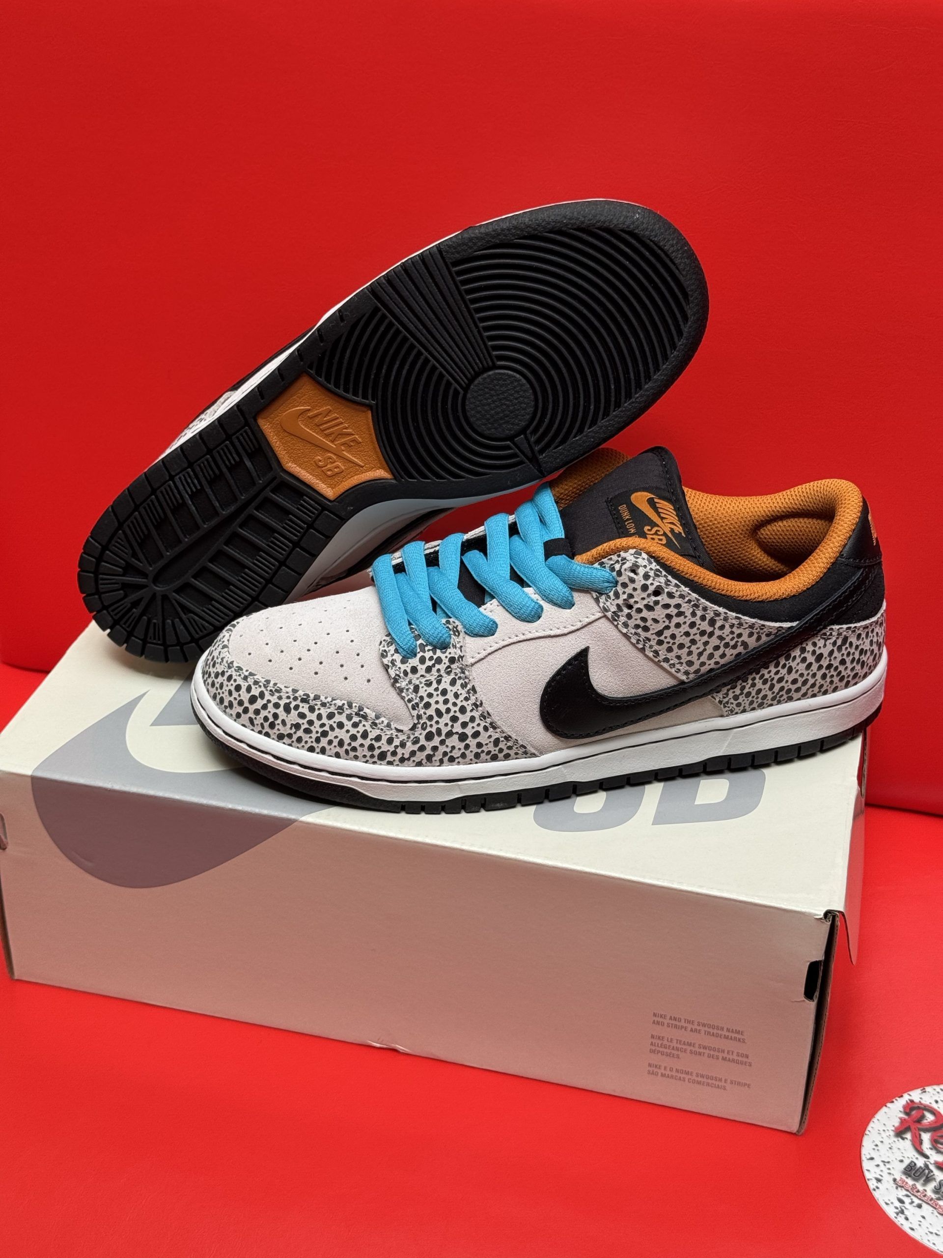Nike SB Dunk Low sneakers, tan with black speckled pattern, blue laces, black sole, on box, red background.