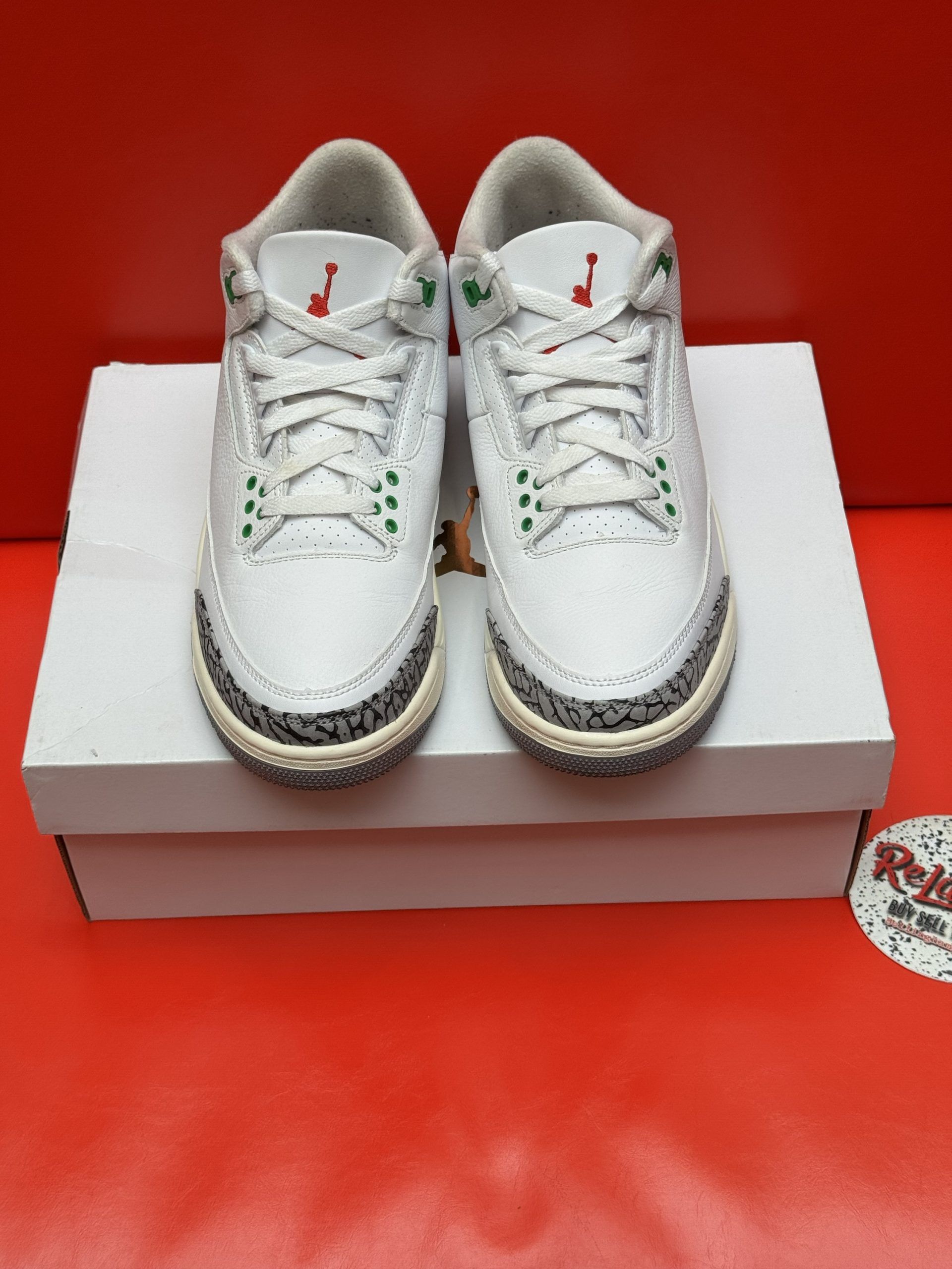 White, green, and gray Air Jordan 3 sneakers on a white box, red background.